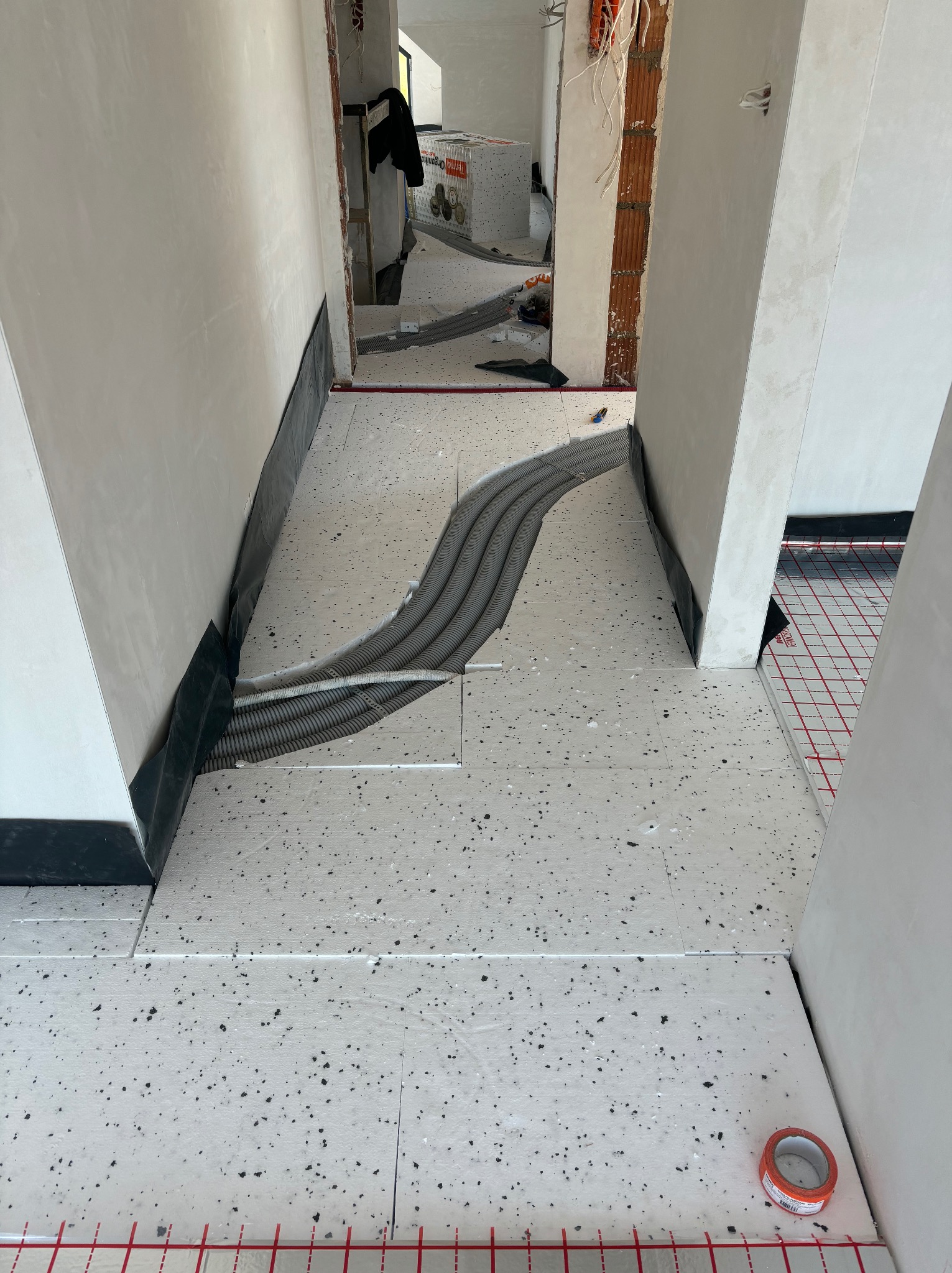 Białe płyty izolacyjne pod ogrzewanie podłogowe z szarymi rurami w bruzdach, widok z góry w trakcie montażu. Czerwona siatka na podłodze w tle.