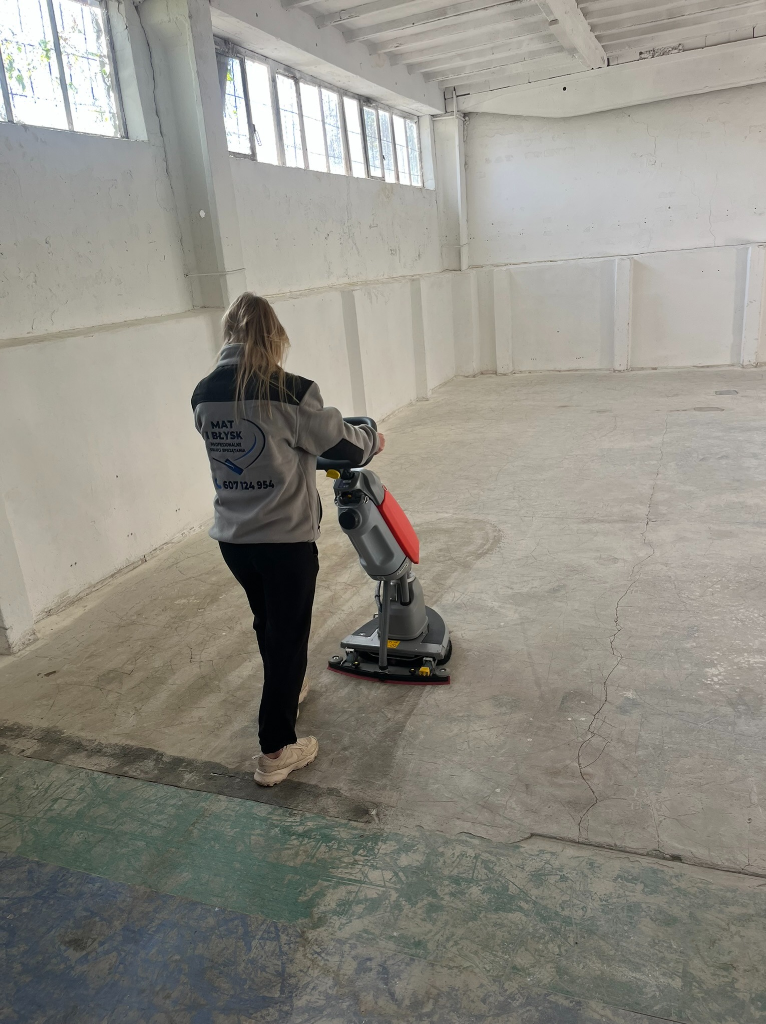 Kobieta w bluzie z logo firmy sprzątającej obsługuje maszynę do czyszczenia posadzki w dużym, surowym pomieszczeniu z betonową podłogą i oknami.