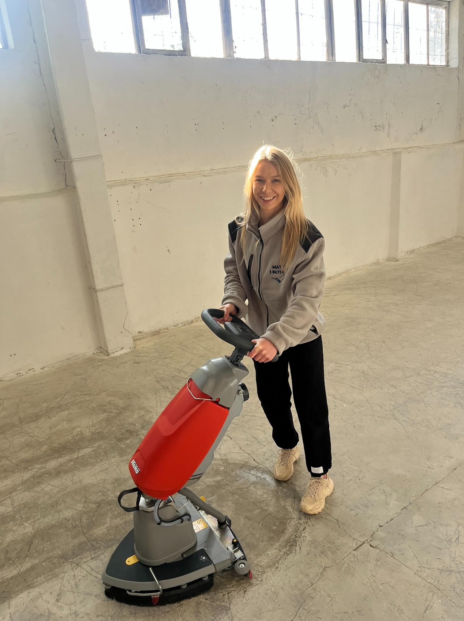 Uśmiechnięta blondynka obsługuje czerwoną maszynę do czyszczenia podłóg Hako w jasnym, surowym wnętrzu z betonową posadzką i oknami w tle.