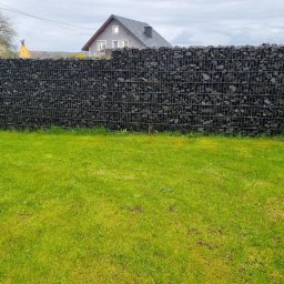 Krak Fence Sp. Z.O.O - Gabionowe ogrodzenie z czarnym wypełnieniem skalnym, widoczne na tle zielonego trawnika i domu z szarą dachówką w pochmurny dzień. Nietypowe wypełnienie koszy gabionowych.