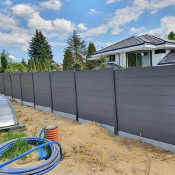 Krak Fence Sp. Z.O.O - Nowoczesne ogrodzenie panelowe w kolorze grafitowym, z widoczną konstrukcją i betonową podmurówką, na tle domu z ogrodem i błękitnego nieba.