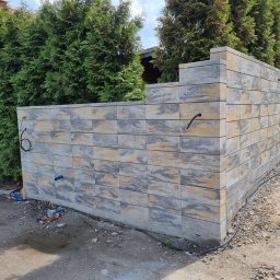 Krak Fence Sp. Z.O.O - Wznoszony mur z szarej, wzorzystej kostki betonowej, tworzący ogrodzenie. Widoczne przewody i narzędzia, sugerujące etap budowy. Tło stanowią zielone tuje i fragment domu.