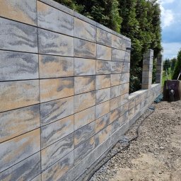 Krak Fence Sp. Z.O.O - Wznoszenie ogrodzenia z dekoracyjnych, dwukolorowych bloczków betonowych. Widoczny fragment muru z przerwą na słupki. W tle zieleń ogrodu i błękitne niebo.