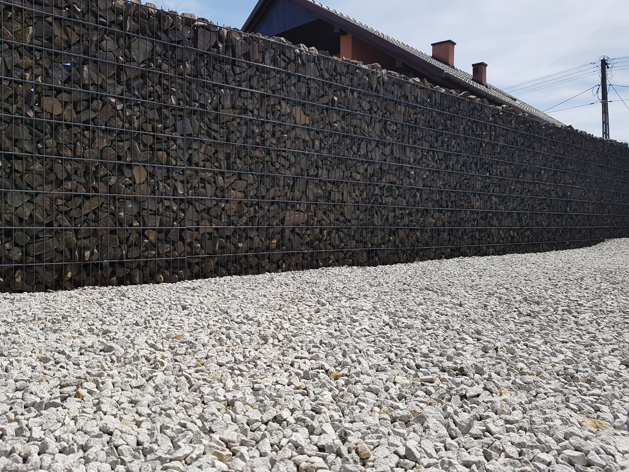 Gabionowe ogrodzenie z ciemnych kamieni, wypełnione w metalowej siatce, z jasnym żwirem u podstawy. W tle widoczny fragment dachu budynku.