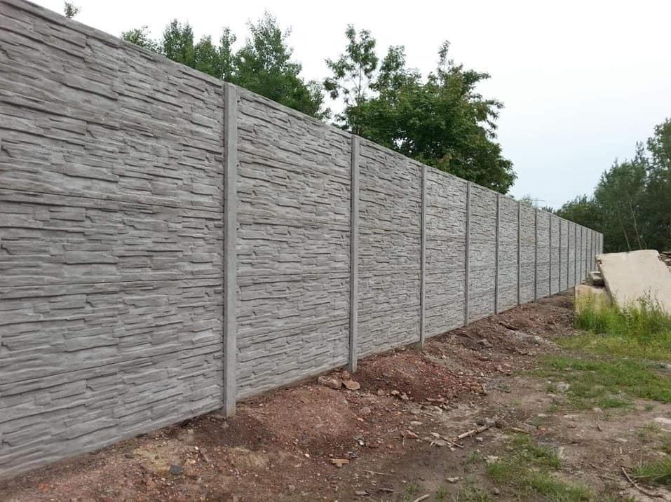 Długi, szary, betonowy płot o teksturze kamienia, z widocznymi słupkami, oddzielający teren. W tle zieleń drzew i krzewów. U dołu ziemia i trawa.