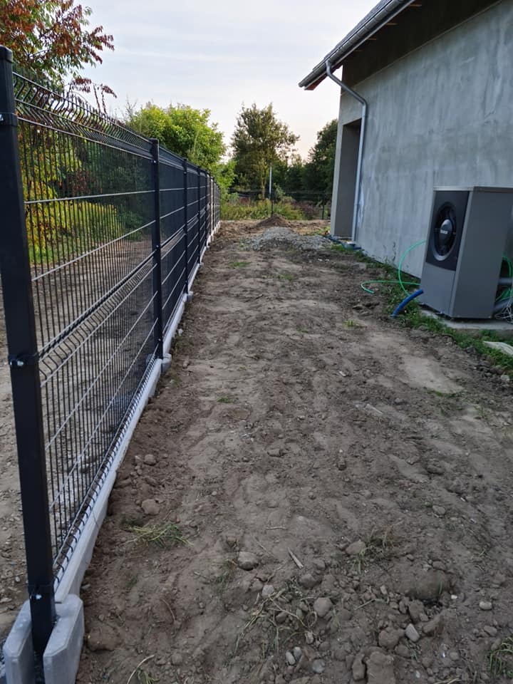 Nowoczesne ogrodzenie panelowe w kolorze grafitowym, z betonową podmurówką, oddzielające posesję od zieleni. Widok z perspektywy gruntu, z budynkiem w tle.