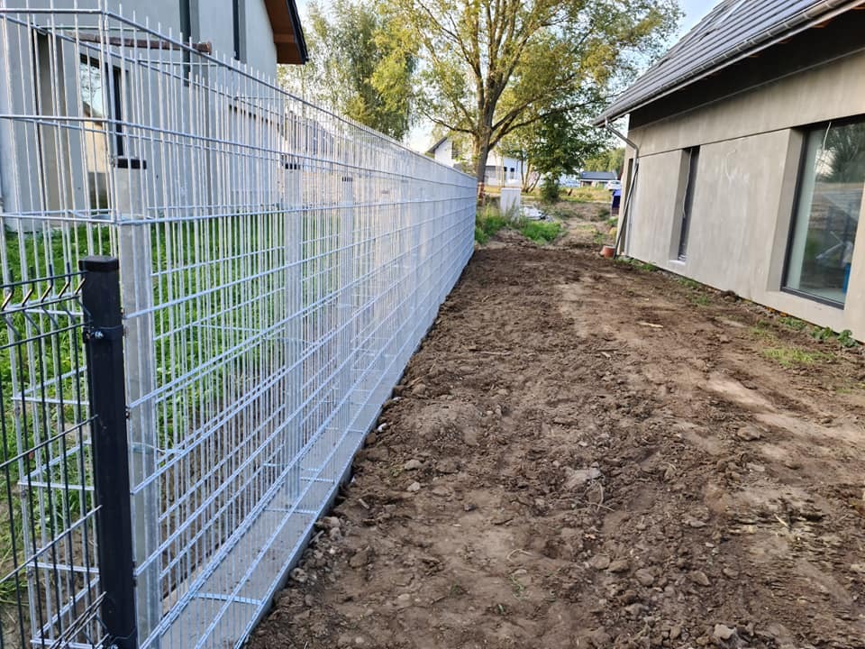 Długi, srebrny panel ogrodzeniowy z czarnymi słupkami, oddzielający posesje. Widok z boku, z ziemią wzdłuż ogrodzenia i budynkami w tle.