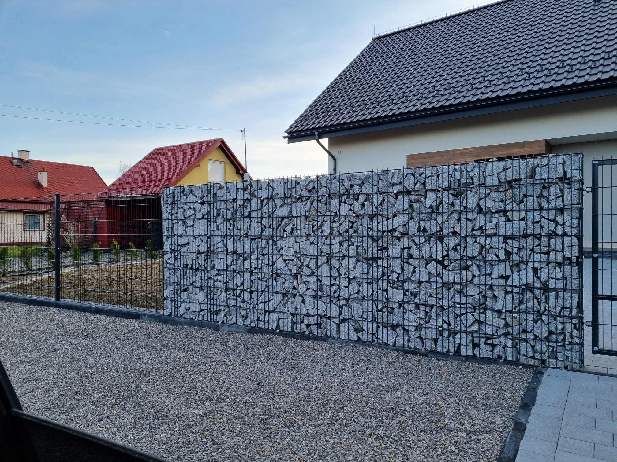 Gabionowe ogrodzenie z szarego kamienia, czarna metalowa siatka, nowoczesny design, na tle domu z ciemną dachówką i żwirowego podłoża. Unikalna tekstura kamienia.