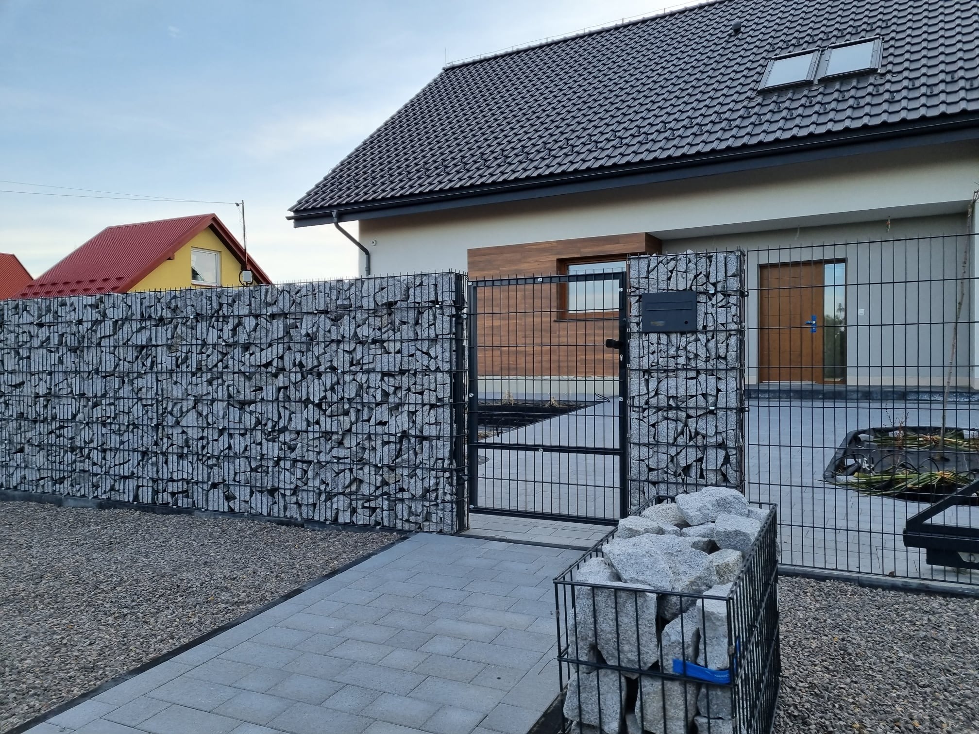 Nowoczesne ogrodzenie gabionowe z metalową bramką wejściową, uzupełnione kamieniami, na tle domu z szarą dachówką i elementami drewnianymi.
