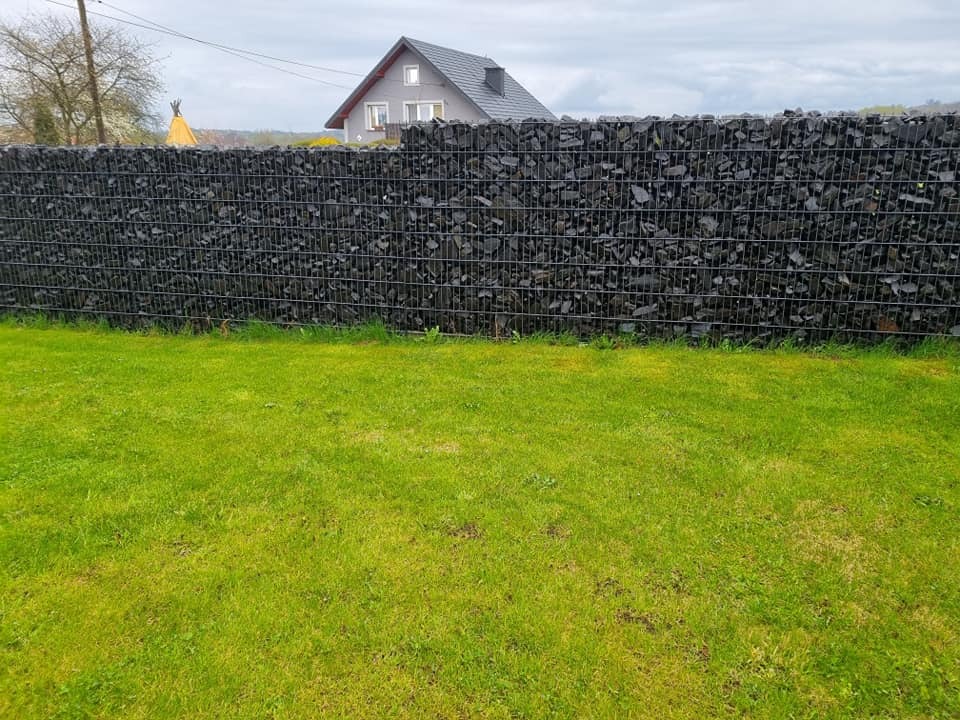 Gabionowe ogrodzenie z czarnym wypełnieniem skalnym, widoczne na tle zielonego trawnika i domu z szarą dachówką w pochmurny dzień. Nietypowe wypełnienie koszy gabionowych.