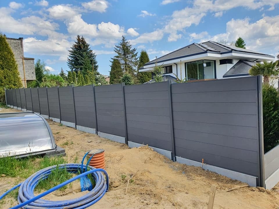 Nowoczesne ogrodzenie panelowe w kolorze grafitowym, z widoczną konstrukcją i betonową podmurówką, na tle domu z ogrodem i błękitnego nieba.