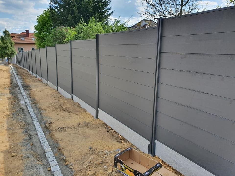 Nowoczesne ogrodzenie panelowe w odcieniach szarości, z widoczną podmurówką i elementami montażowymi. Ujęcie z perspektywy ziemi, podkreślające długość i solidność konstrukcji.