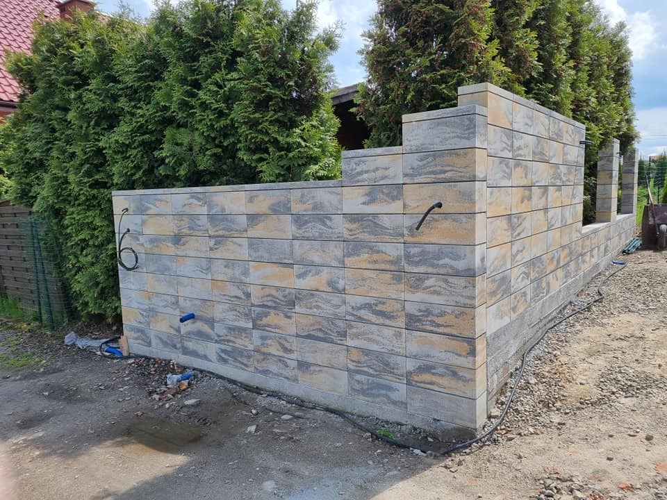 Wznoszony mur z szarej, wzorzystej kostki betonowej, tworzący ogrodzenie. Widoczne przewody i narzędzia, sugerujące etap budowy. Tło stanowią zielone tuje i fragment domu.