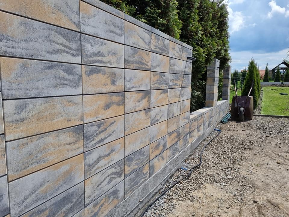 Wznoszenie ogrodzenia z dekoracyjnych, dwukolorowych bloczków betonowych. Widoczny fragment muru z przerwą na słupki. W tle zieleń ogrodu i błękitne niebo.
