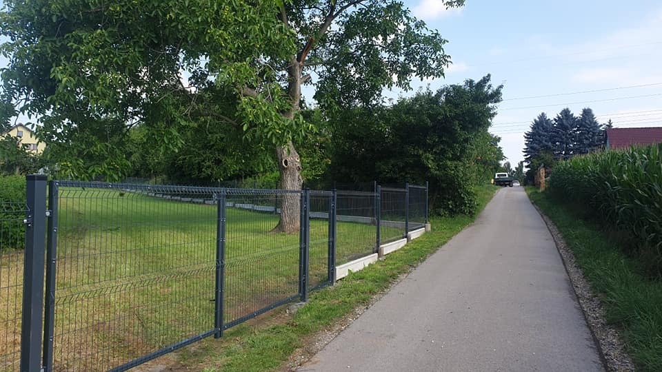 Nowoczesne ogrodzenie panelowe w kolorze grafitowym, z betonową podmurówką, oddzielające posesję od drogi. W tle zieleń drzew i krzewów, a w oddali zabudowania.