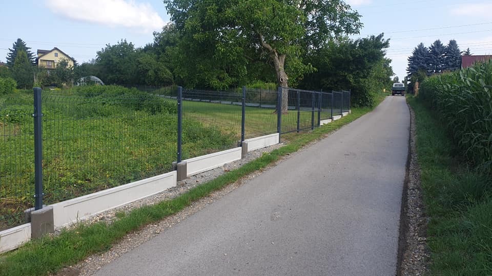 Długi, szary metalowy płot panelowy z betonową podmurówką, biegnący wzdłuż asfaltowej drogi wiejskiej, otoczony zielenią i drzewami w słoneczny dzień.