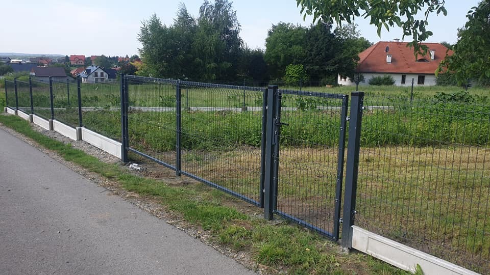 Nowoczesne ogrodzenie panelowe z bramką, szare słupki, betonowa podmurówka, widok z zewnątrz na tle zieleni i zabudowań.