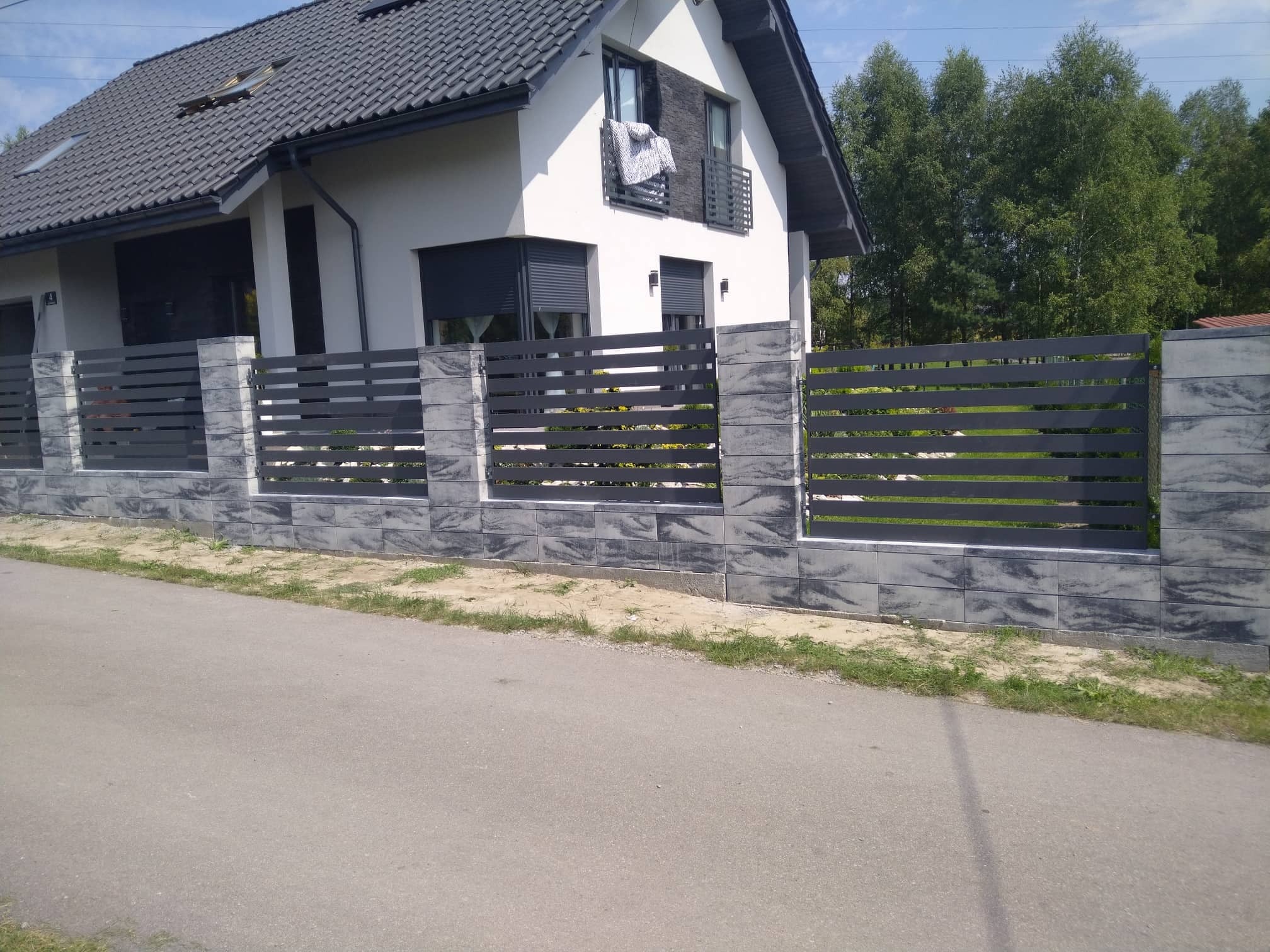 Nowoczesne ogrodzenie posesji z szarego, poziomego panelu i betonowych słupków z fakturą kamienia, na tle białego domu z ciemną dachówką i zieleni drzew.