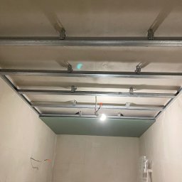 Paweł Orzechowski - Montaż sufitu podwieszanego: metalowa konstrukcja z częściowo zamontowaną zieloną płytą. Widoczne przewody elektryczne i drabina. Wnętrze w trakcie prac wykończeniowych.