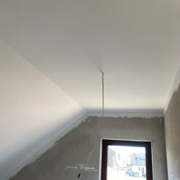 Paweł Orzechowski - Biały sufit pod skosem po malowaniu, widoczne ślady szpachlowania na ścianach, okno z ciemną ramą. Kabel zwisa z sufitu. Wnętrze w trakcie wykańczania.
