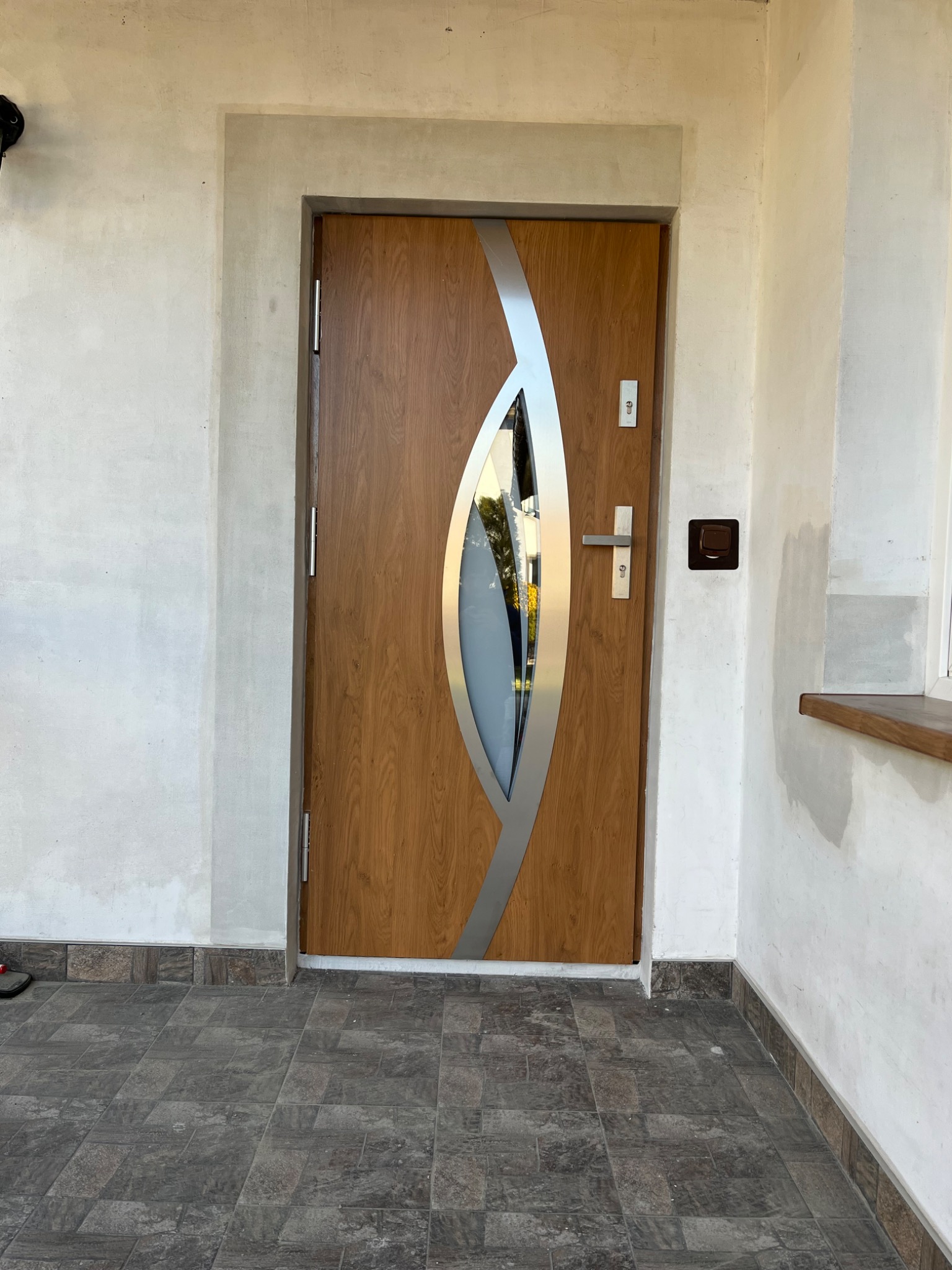 Brązowe drzwi wejściowe z szybą i srebrnymi zdobieniami, osadzone w surowej, nieotynkowanej wnęce. Widoczne płytki podłogowe i fragment ściany z przełącznikiem.