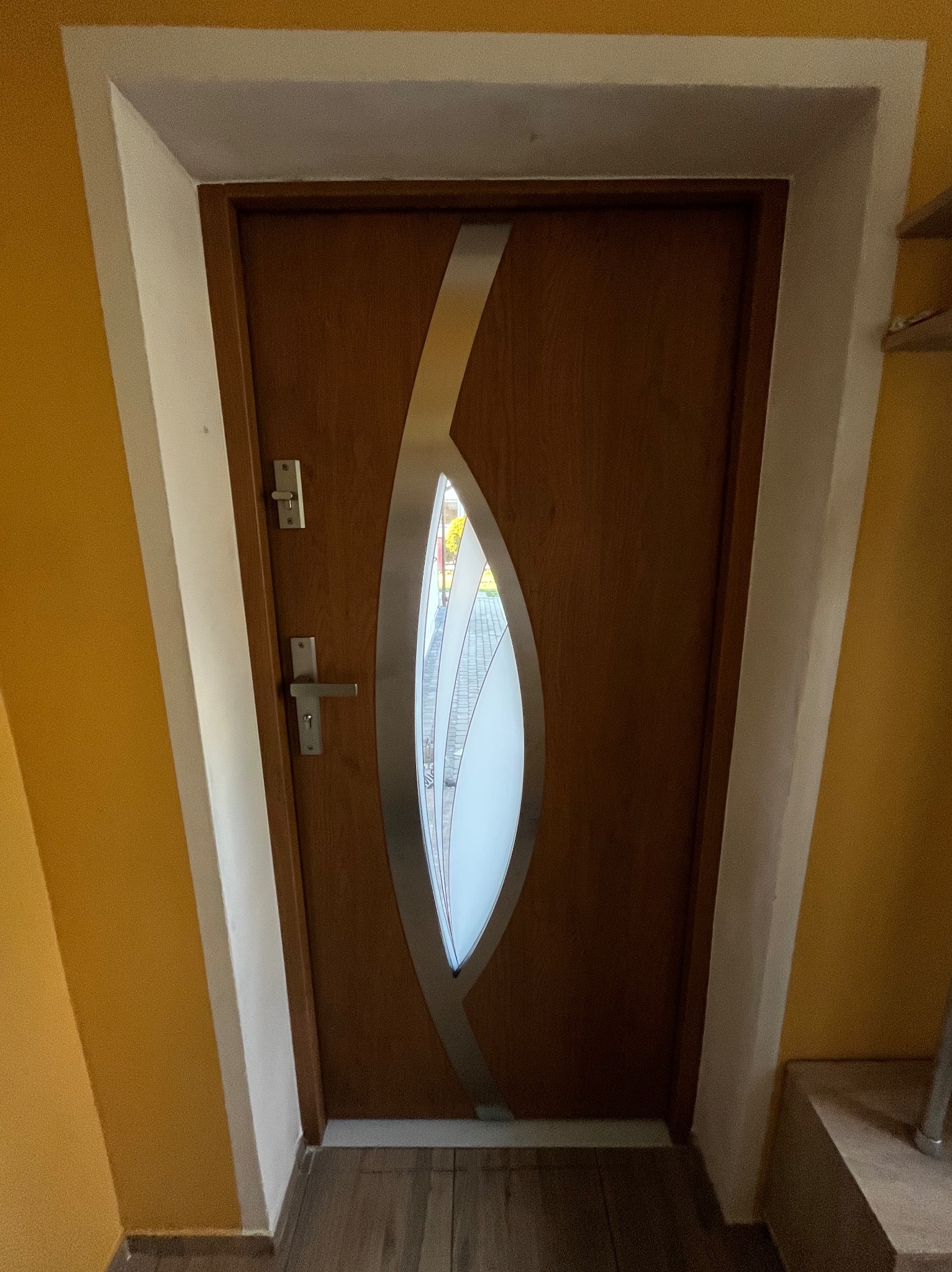 Brązowe drzwi wejściowe z owalnym przeszkleniem i srebrną aplikacją, osadzone w białej ościeżnicy na tle żółtej ściany. Widoczny fragment podłogi i schodów.