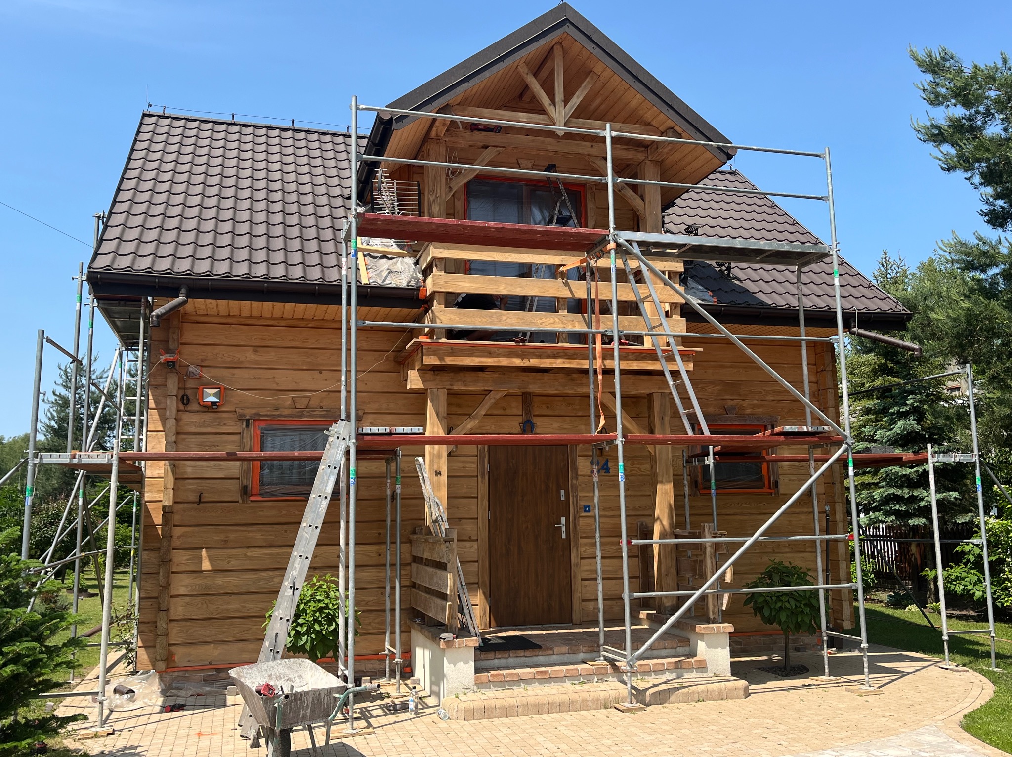 Drewniany dom w trakcie budowy/renowacji, otoczony rusztowaniami. Widoczne elementy konstrukcyjne, okna zabezpieczone folią, wózek budowlany. Słoneczny dzień.