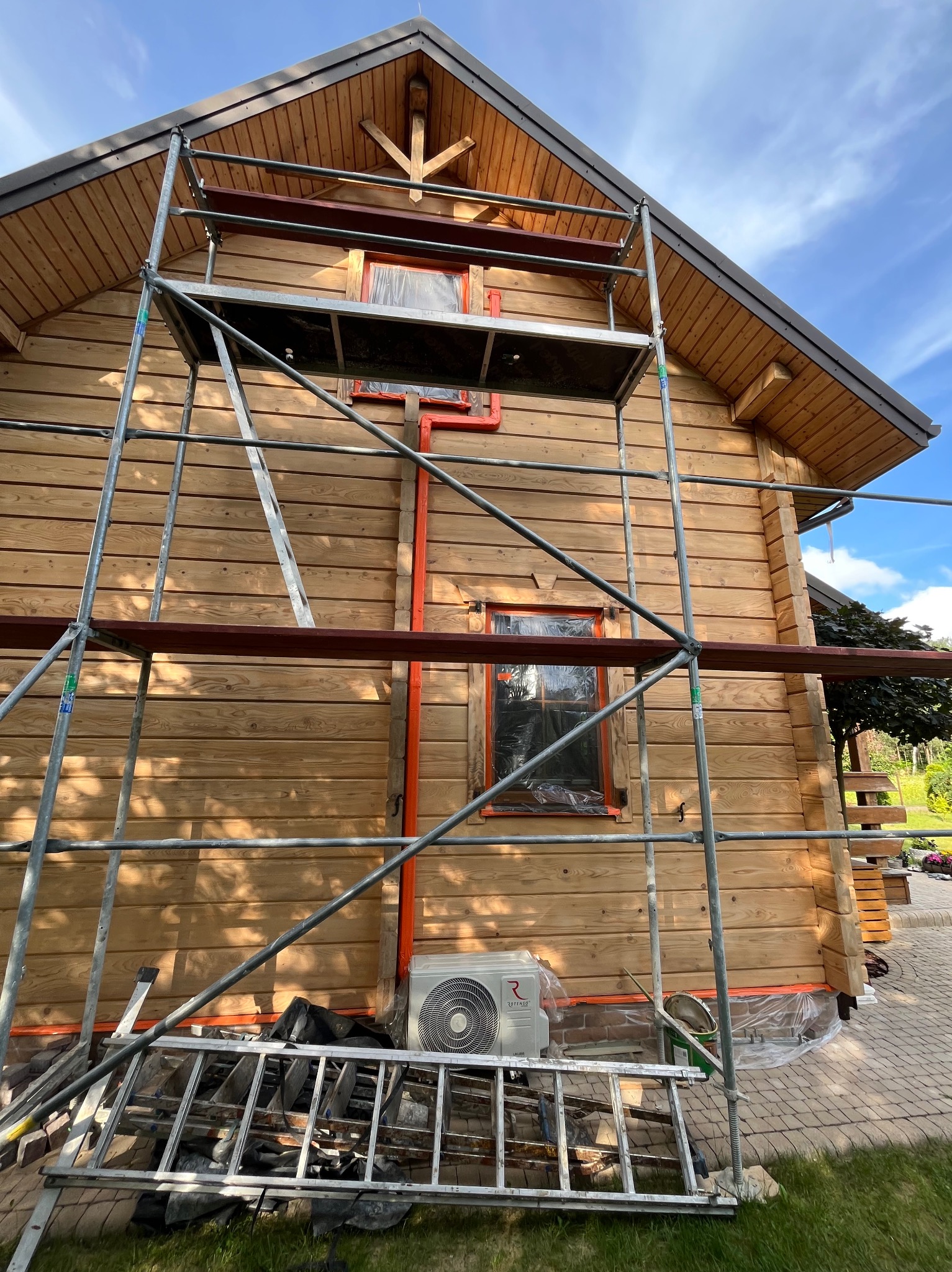 Drewniany dom w trakcie prac wykończeniowych, z rusztowaniem i zabezpieczonymi oknami. Widoczny klimatyzator i drabina, sugerujące prace zewnętrzne.