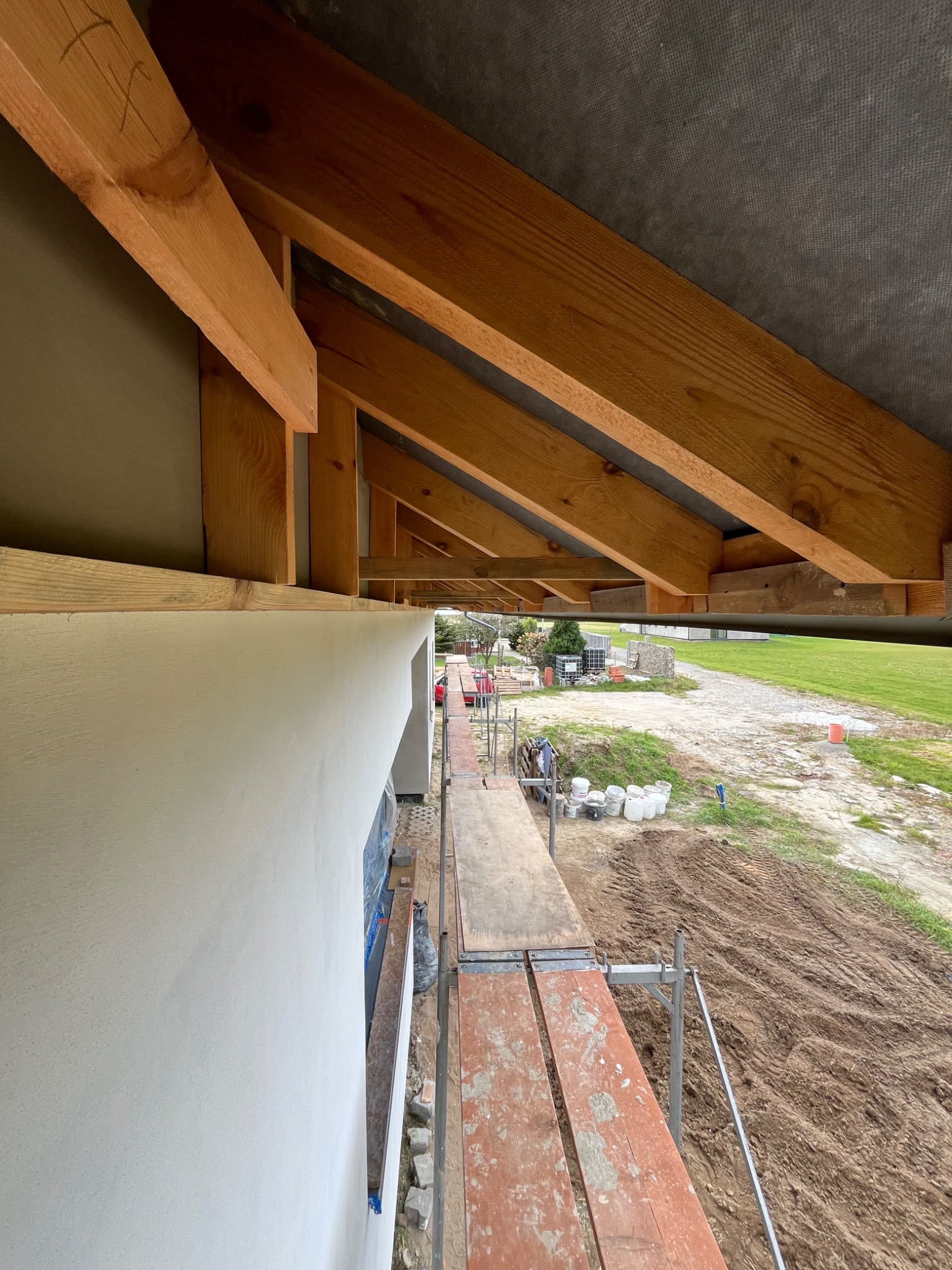 Ocieplony budynek z drewnianą konstrukcją dachu, widok z rusztowania. Świeżo otynkowana ściana zewnętrzna, widoczne elementy więźby dachowej i otoczenie budynku.