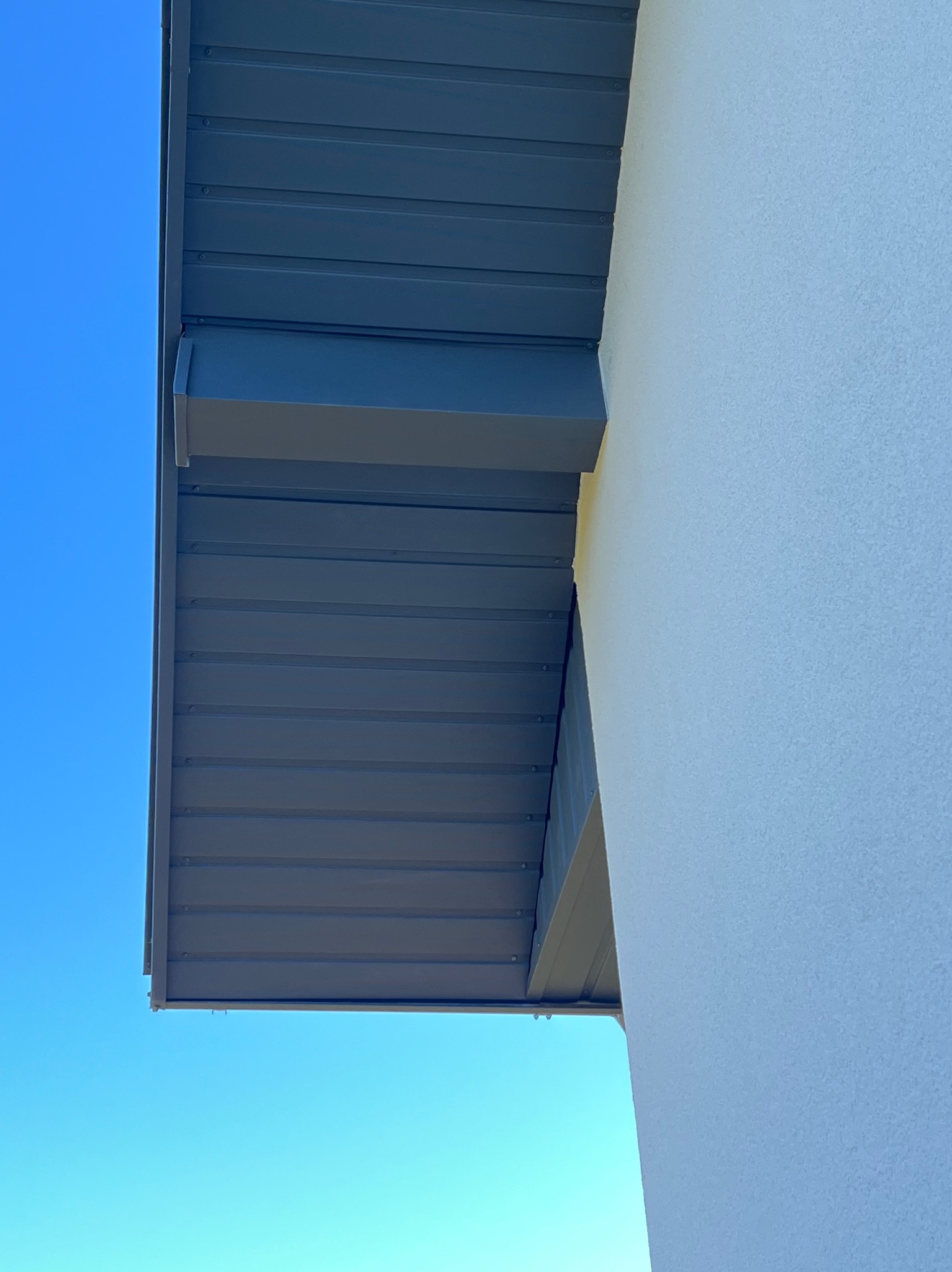 Detale wykończenia elewacji budynku: szara, metalowa podbitka dachowa z widocznymi mocowaniami i fragment ściany o jasnej, chropowatej fakturze na tle błękitnego nieba.