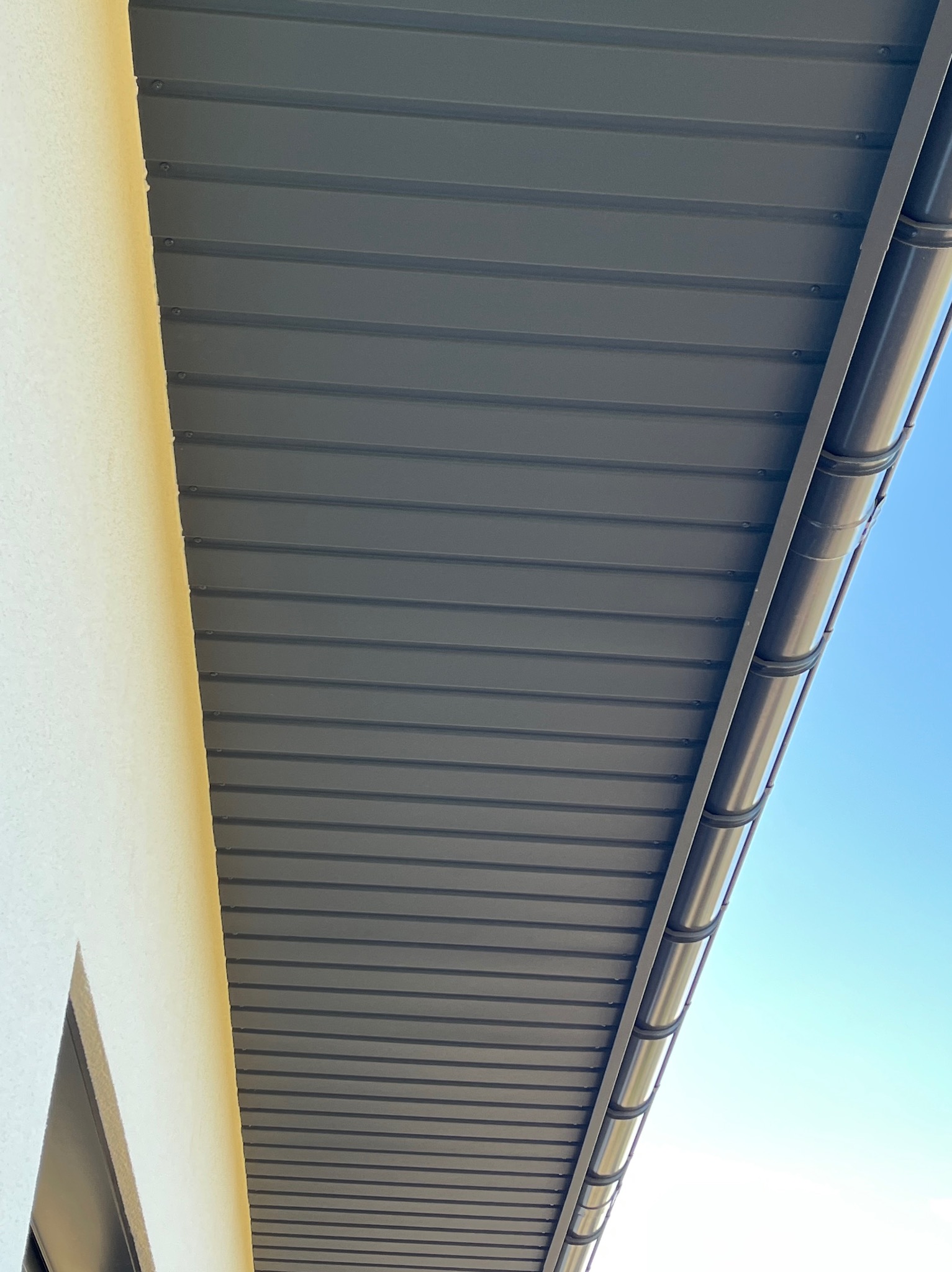 Szare podbitka dachowa z widocznym systemem rynnowym, zamontowana przy elewacji budynku, na tle błękitnego nieba. Ujęcie z dołu, podkreślające detale montażu.