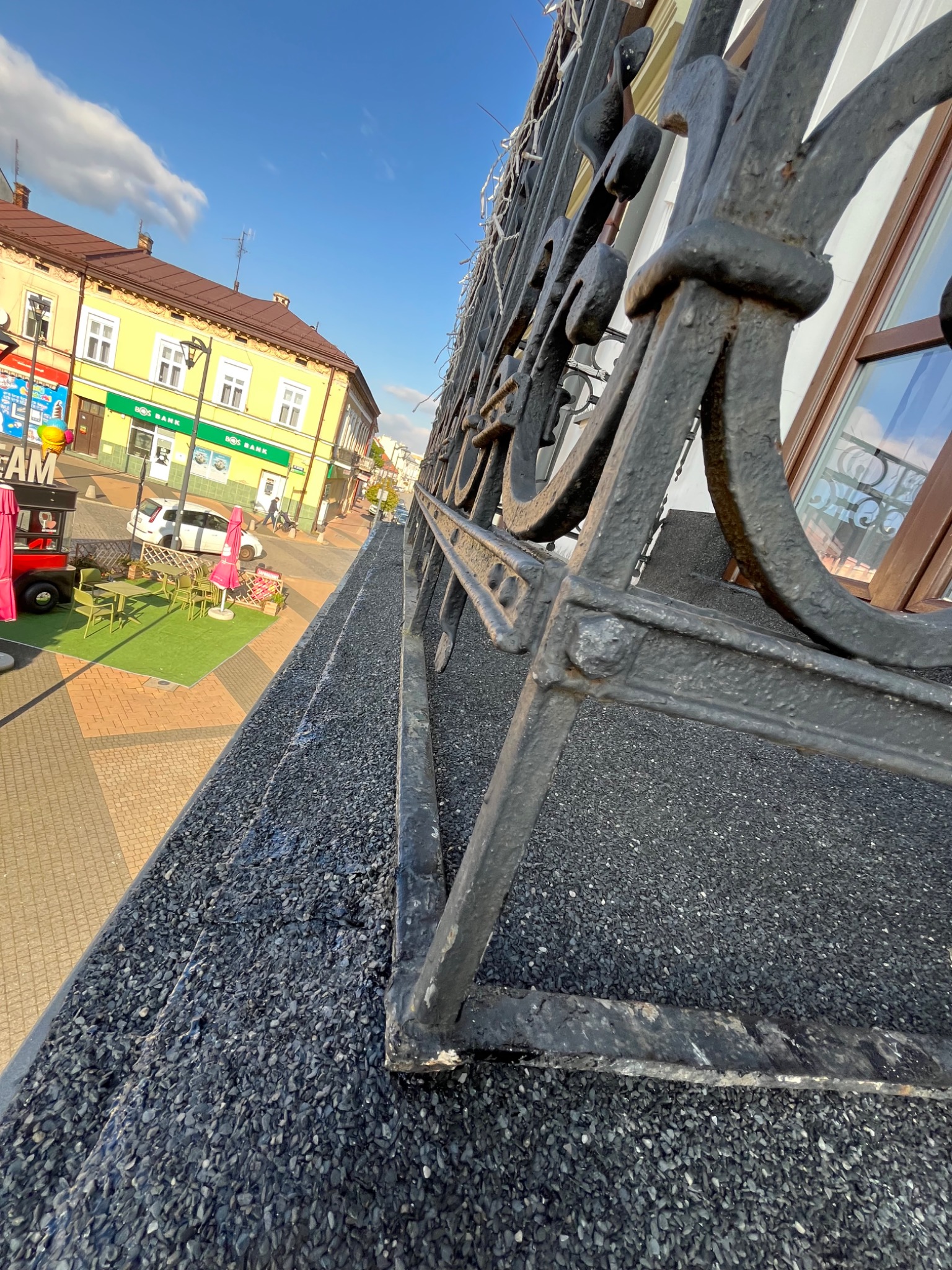 Kuty balkon z widokiem na miasto w słoneczny dzień. Balustrada z metalu, tło stanowi budynek z oknami i fragment ulicy z elementami małej architektury.