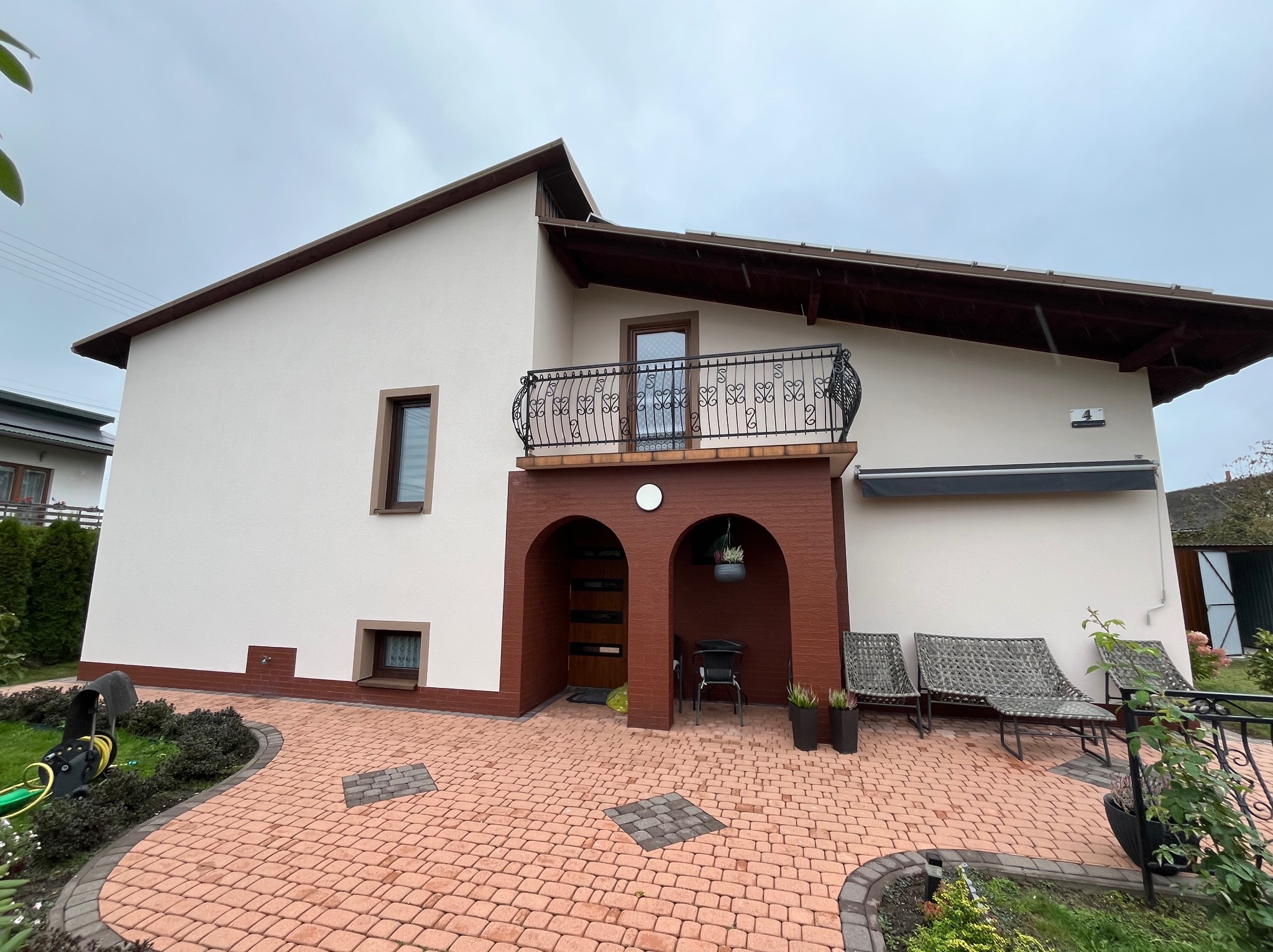 Elewacja domu jednorodzinnego po renowacji, z balkonem z ozdobną balustradą i nową kostką brukową na podjeździe. Widoczne detale architektoniczne i staranne wykończenie.