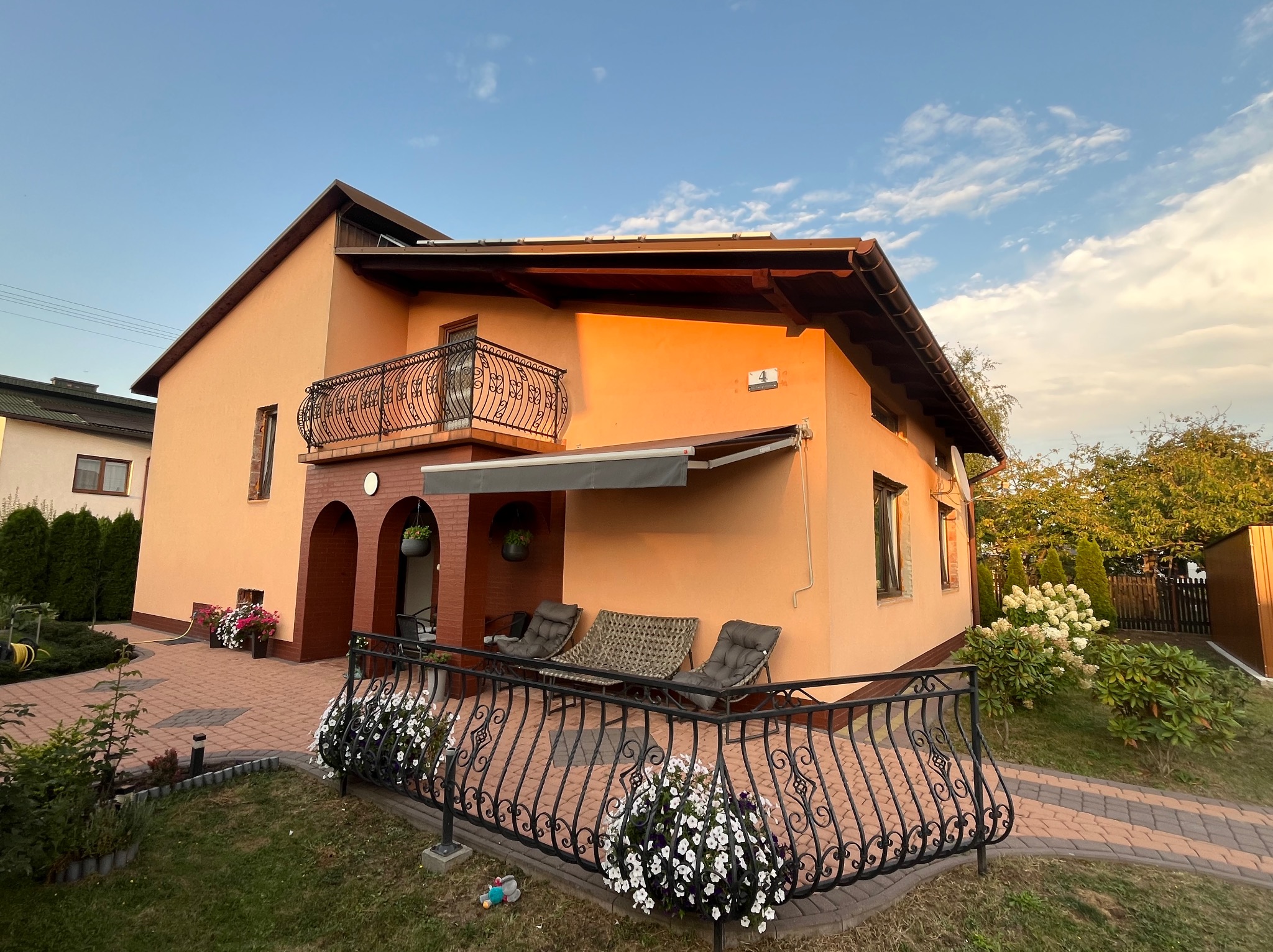 Elewacja domu jednorodzinnego w ciepłych barwach, z balkonem z kutą balustradą i markizą. Widoczne elementy dekoracyjne i zadbany ogród.