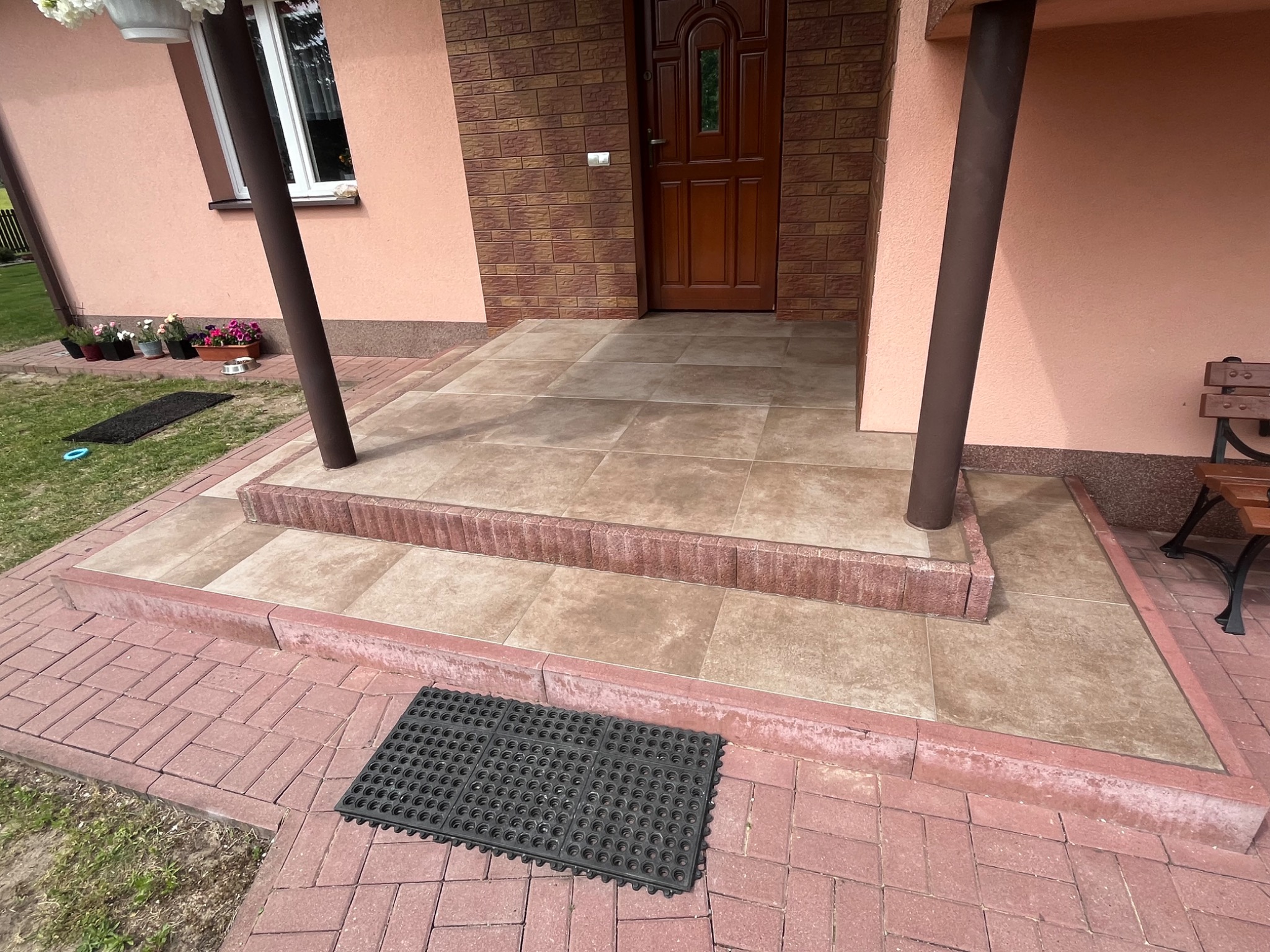 Wykończone płytkami schody wejściowe do domu z brązowymi kolumnami i ceglaną elewacją. Ciemna wycieraczka leży na czerwonej kostce brukowej.
