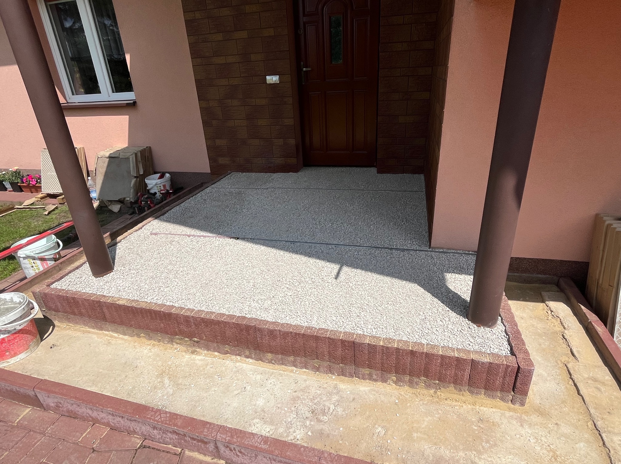 Wykończone wejście do domu z nową, jasną nawierzchnią z drobnych kamyczków, obramowane ceglaną kostką. Widoczne filary podtrzymujące zadaszenie i fragment elewacji.