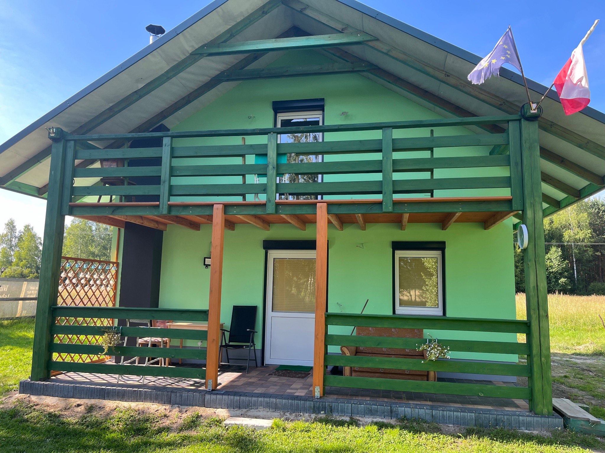Zielony dom z drewnianym balkonem i werandą, otoczony zielenią. Flagi Polski i UE powiewają na wietrze. Widoczne meble ogrodowe i elementy dekoracyjne.