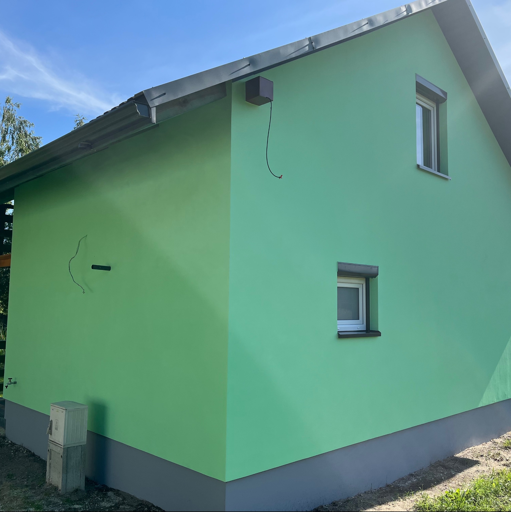 Ściana budynku po malowaniu na zielono w Hucina, widoczne dwa okna i skrzynka elektryczna. Świeży kolor elewacji, gładka powierzchnia.