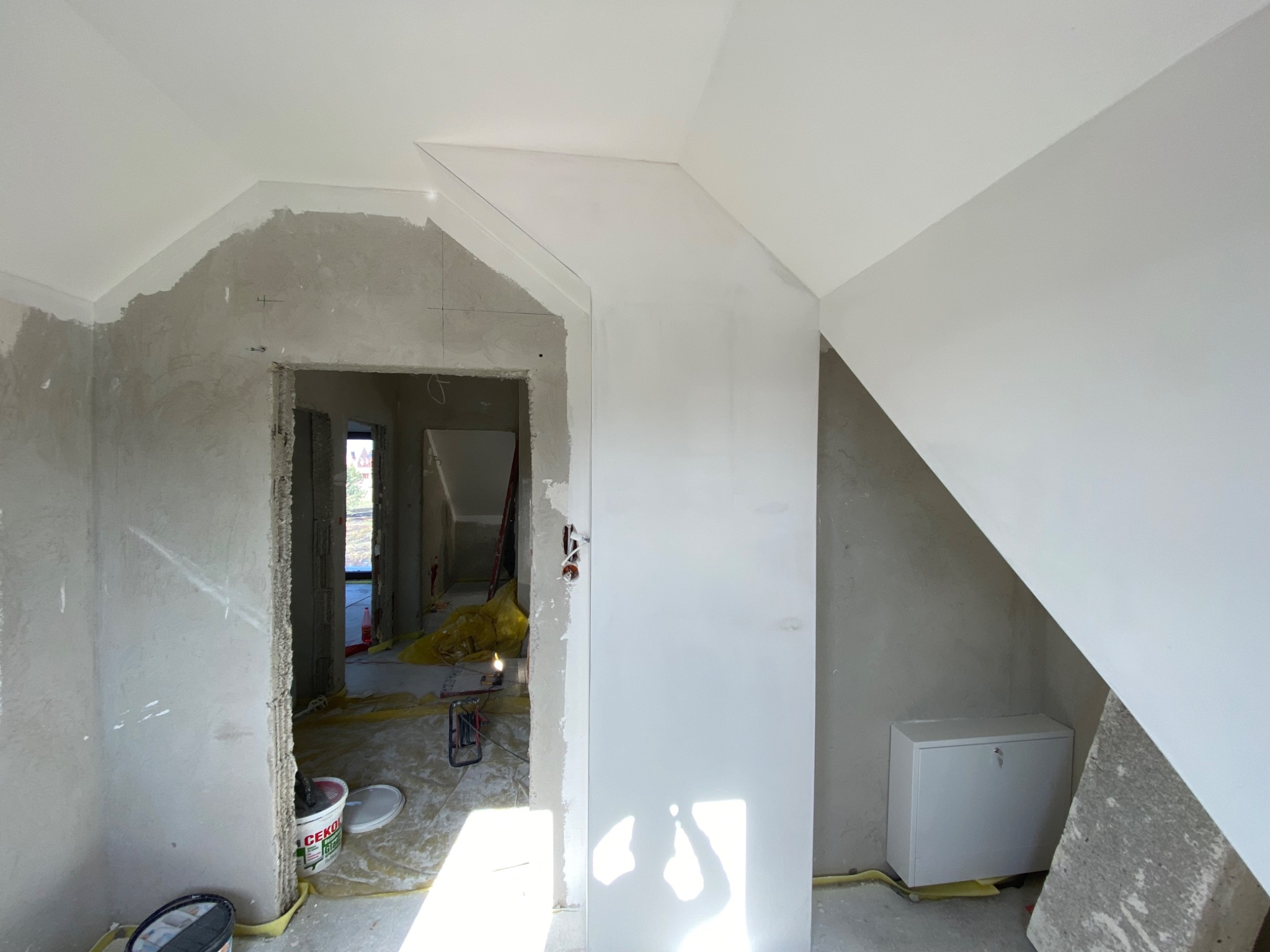 Wnętrze poddasza w trakcie prac wykończeniowych: ściany częściowo wyszpachlowane, widoczne narzędzia i materiały budowlane, surowe oświetlenie.