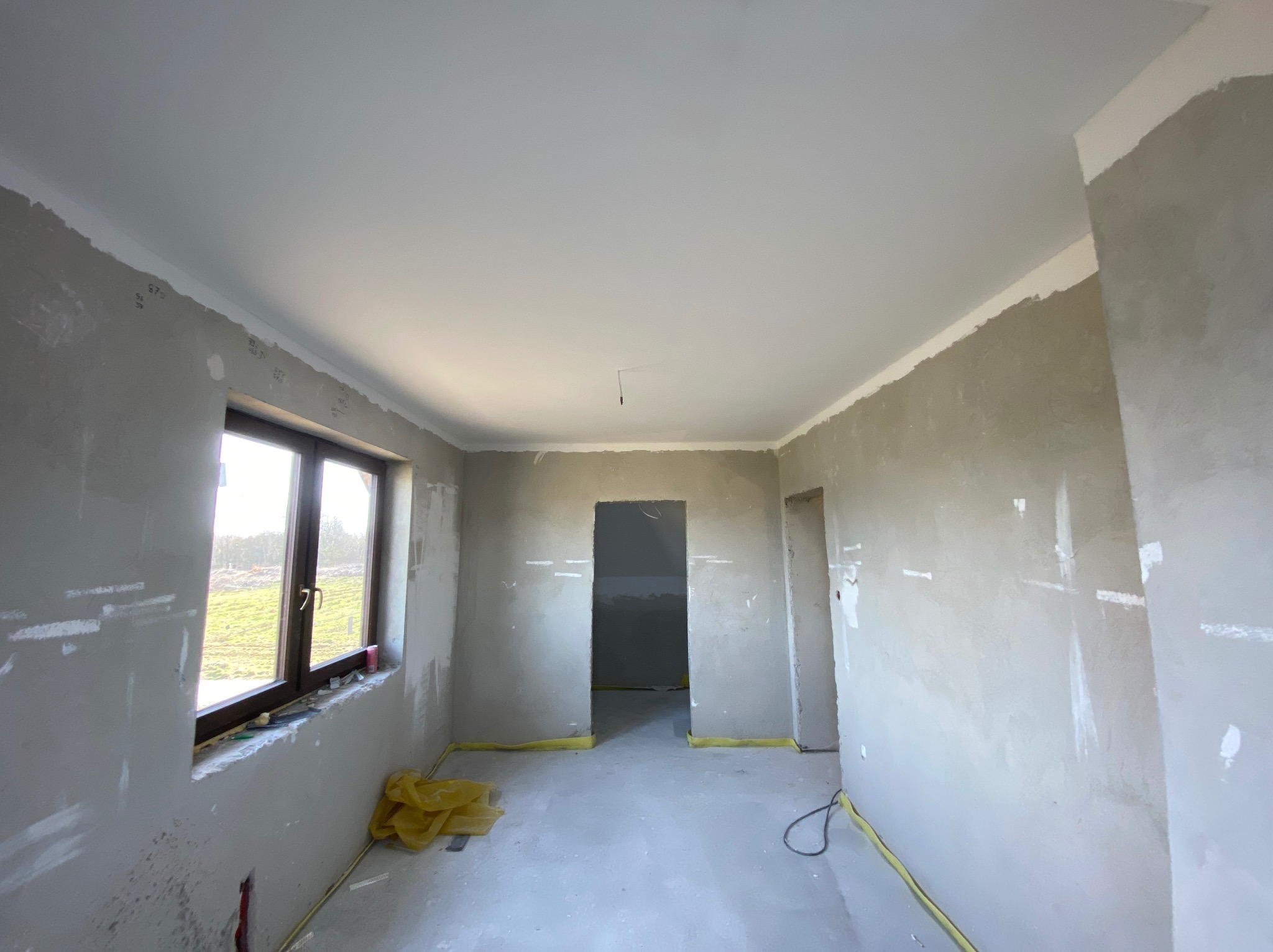 Wnętrze w trakcie szpachlowania: ściany pokryte szpachlą, okno z widokiem na zewnątrz, zabezpieczona podłoga. Widoczne ślady po łatach i przygotowaniu do malowania.