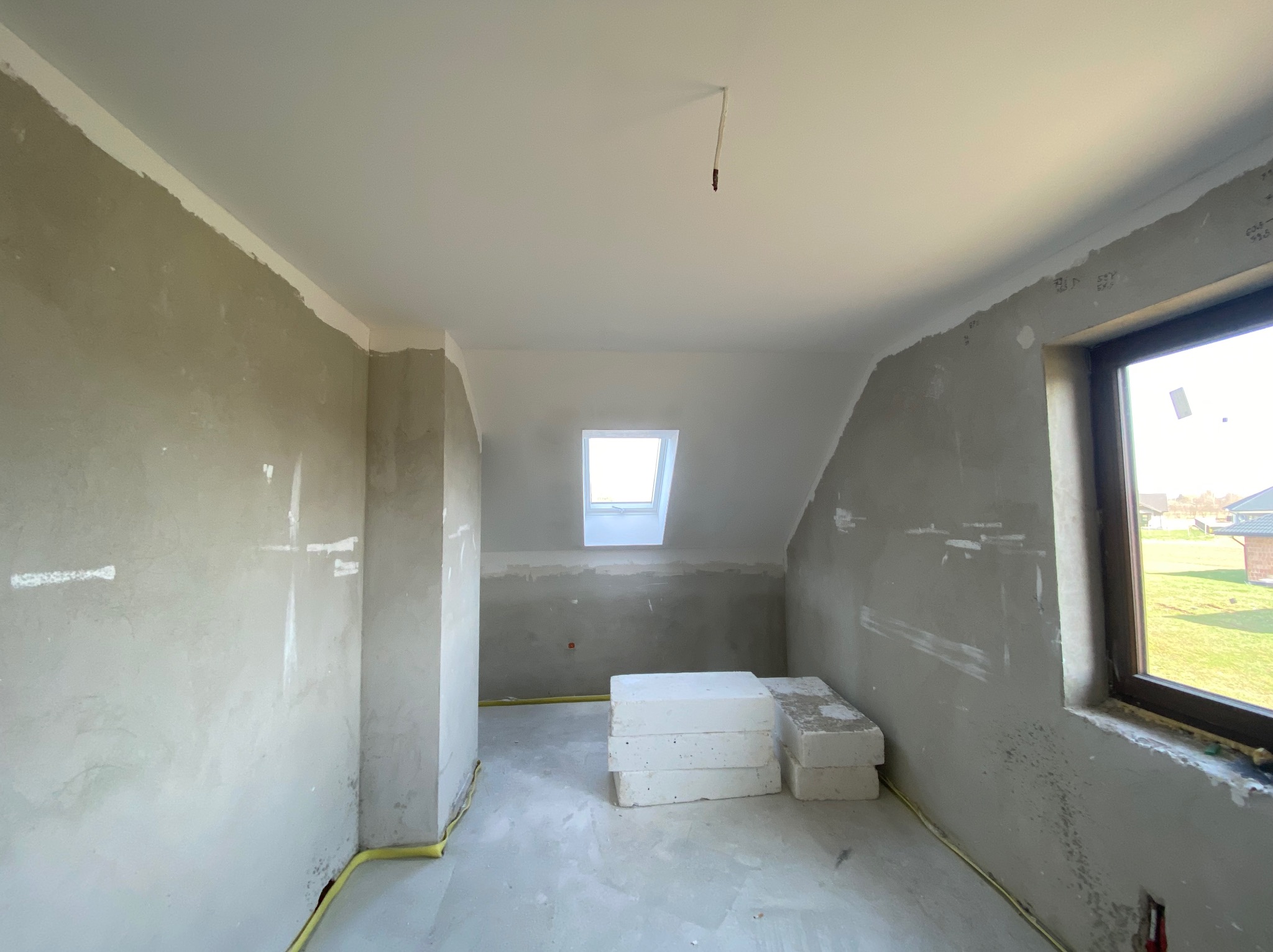 Poddasze w trakcie szpachlowania z oknem dachowym i widocznymi przewodami elektrycznymi przy podłodze. Ściany przygotowane do malowania, widoczne bloczki styropianowe.