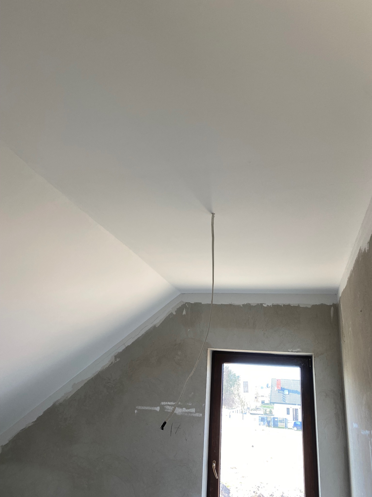 Biały sufit pod skosem po malowaniu, widoczne ślady szpachlowania na ścianach, okno z ciemną ramą. Kabel zwisa z sufitu. Wnętrze w trakcie wykańczania.