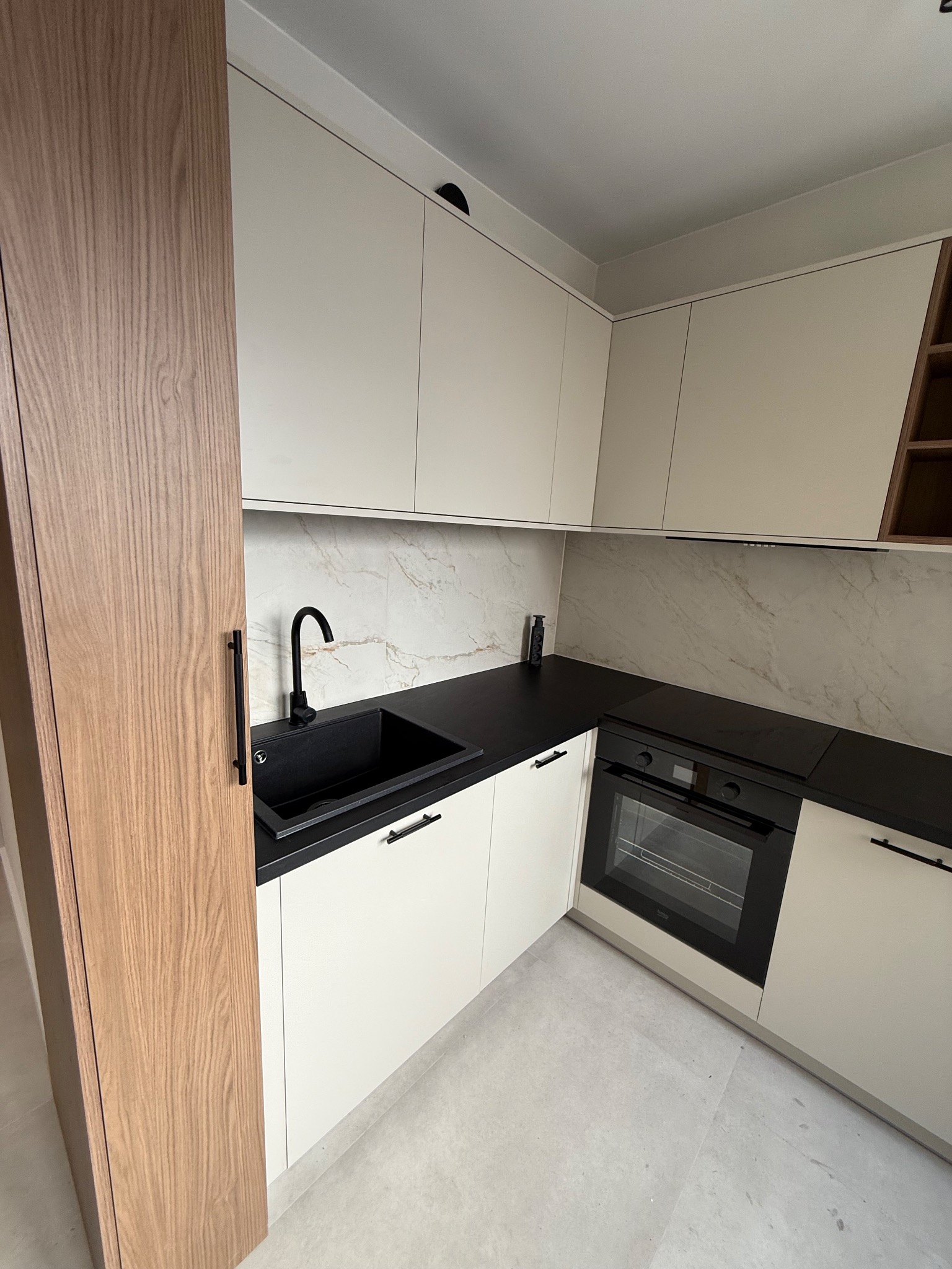 Jasna kuchnia z czarnym blatem i zlewozmywakiem, fronty szafek bez uchwytów, marmurowe wykończenie ściany nad blatem, drewniana szafka po lewej stronie.