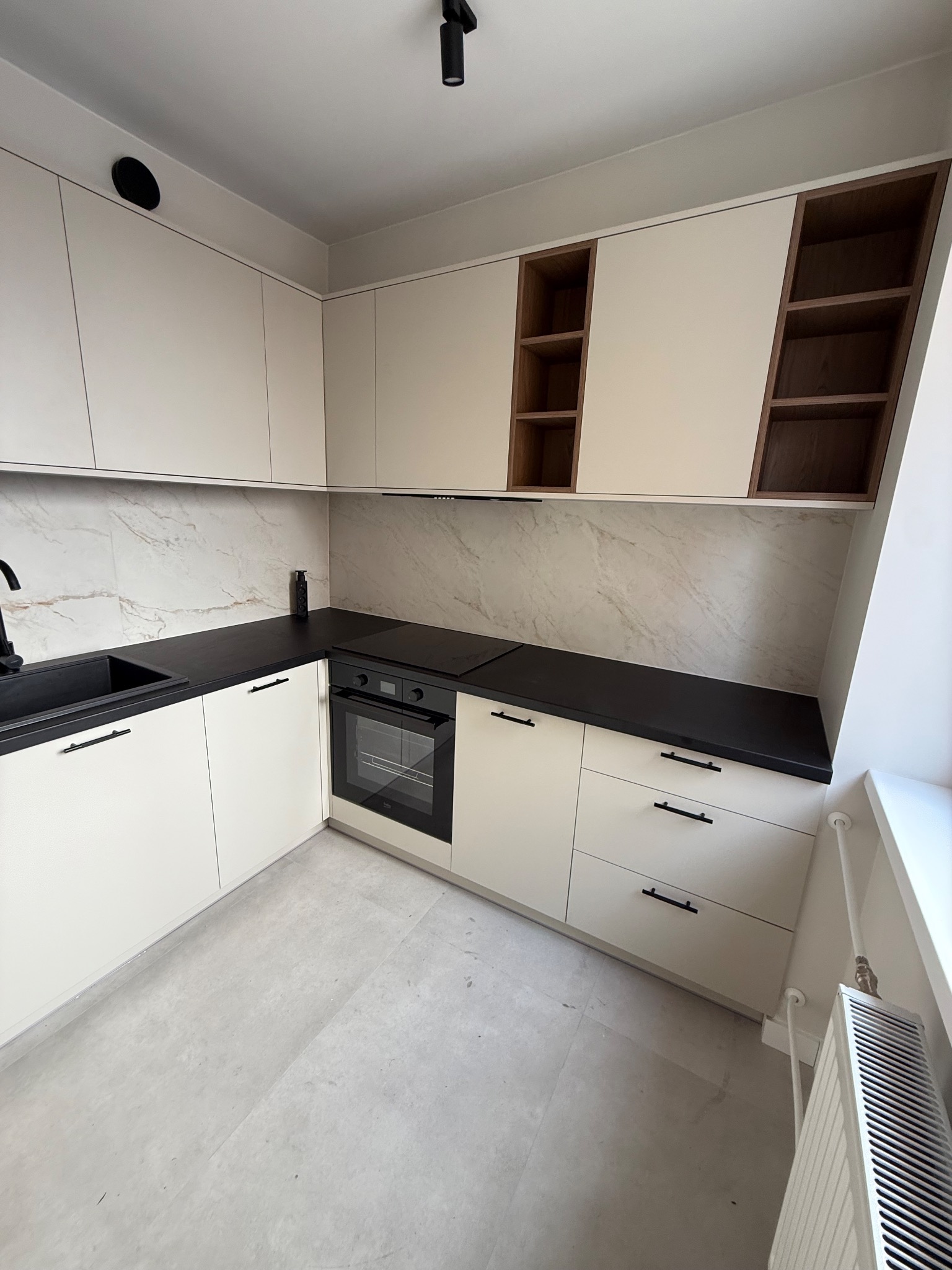 Jasna kuchnia z czarnym blatem i elementami drewna. Uwagę przykuwają otwarte półki w górnych szafkach i minimalistyczne, czarne uchwyty. Nowoczesny design.