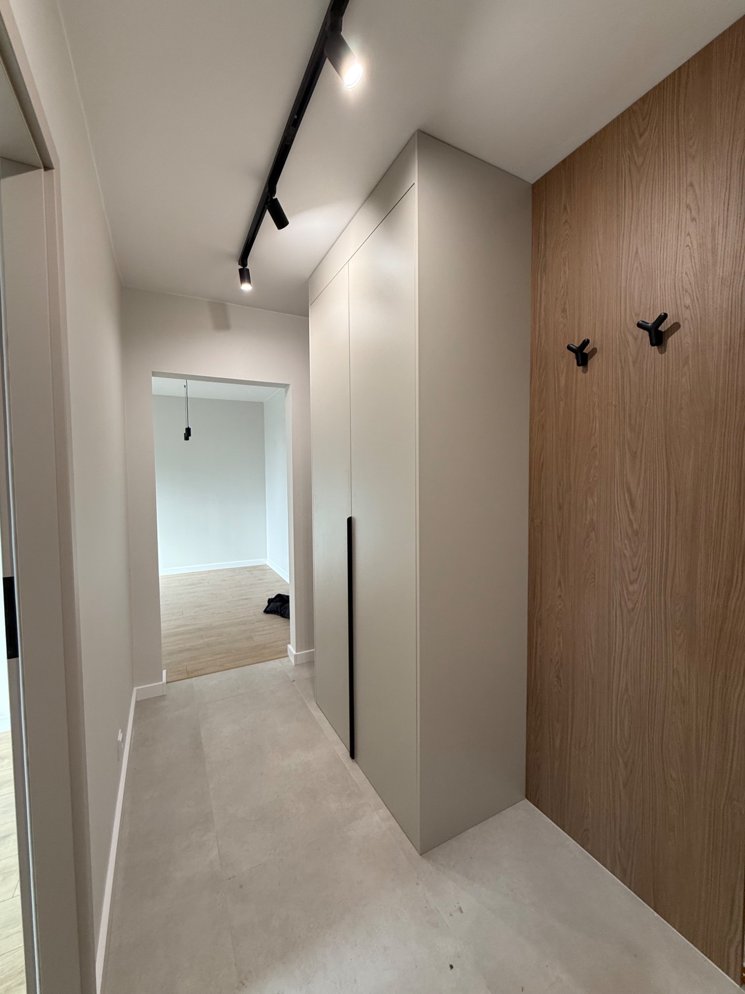 Nowoczesny przedpokój z szafą w zabudowie, drewnianą ścianą z wieszakami i szynoprzewodem oświetleniowym. Minimalistyczny design, neutralna kolorystyka.