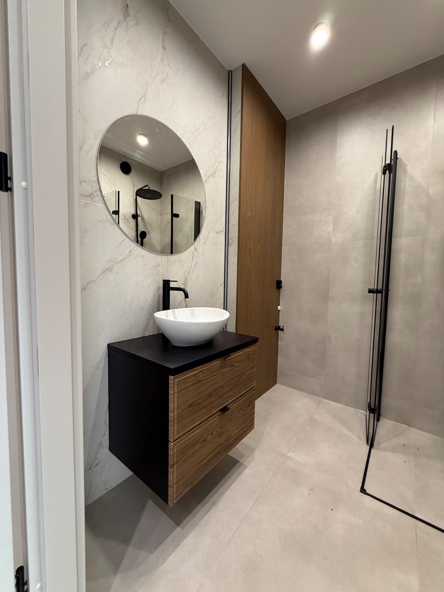 Nowoczesna łazienka z szafką pod umywalkę w kolorze czarnym i drewnopodobnym, okrągłe lustro, armatura w czarnym macie i kabina prysznicowa typu walk-in.