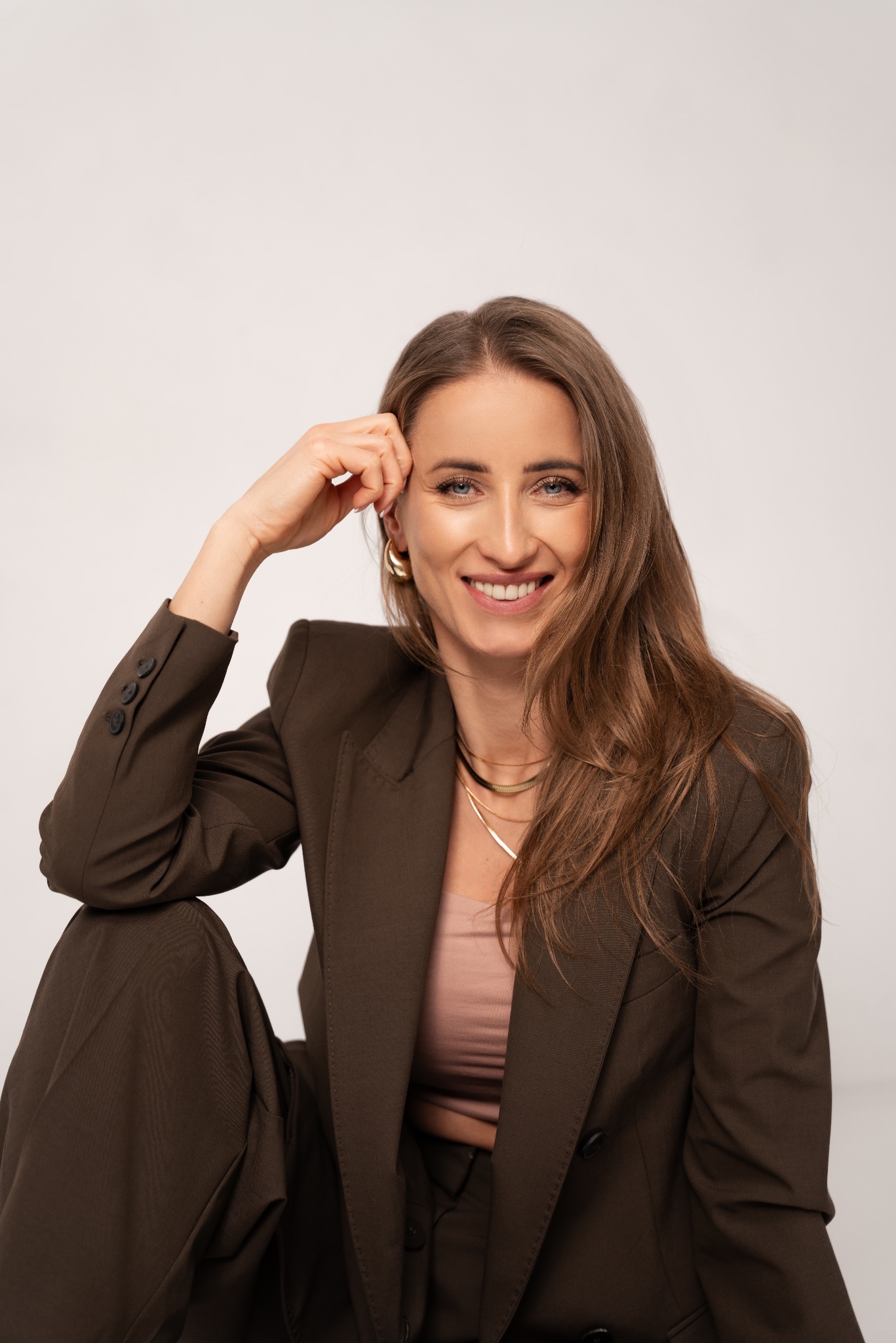 Uśmiechnięta agentka nieruchomości w brązowym garniturze, z ręką przy głowie, na jasnym tle. Profesjonalny portret biznesowy z delikatnym makijażem i złotą biżuterią.