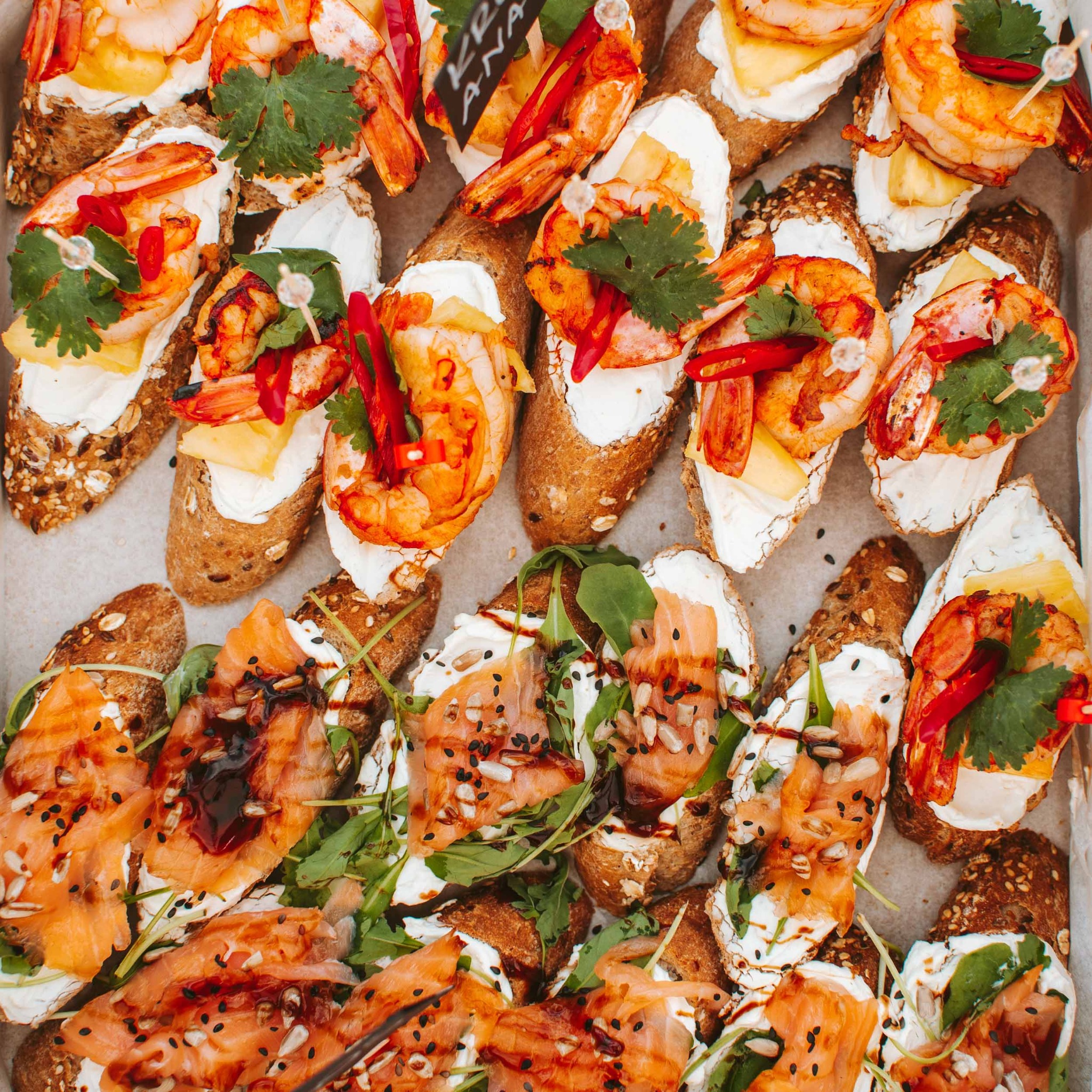 Różnorodne kanapki cateringowe na ciemnym pieczywie: krewetki z ananasem i chili, łosoś z rukolą i sosem balsamicznym, ułożone w kartonowym pudełku.