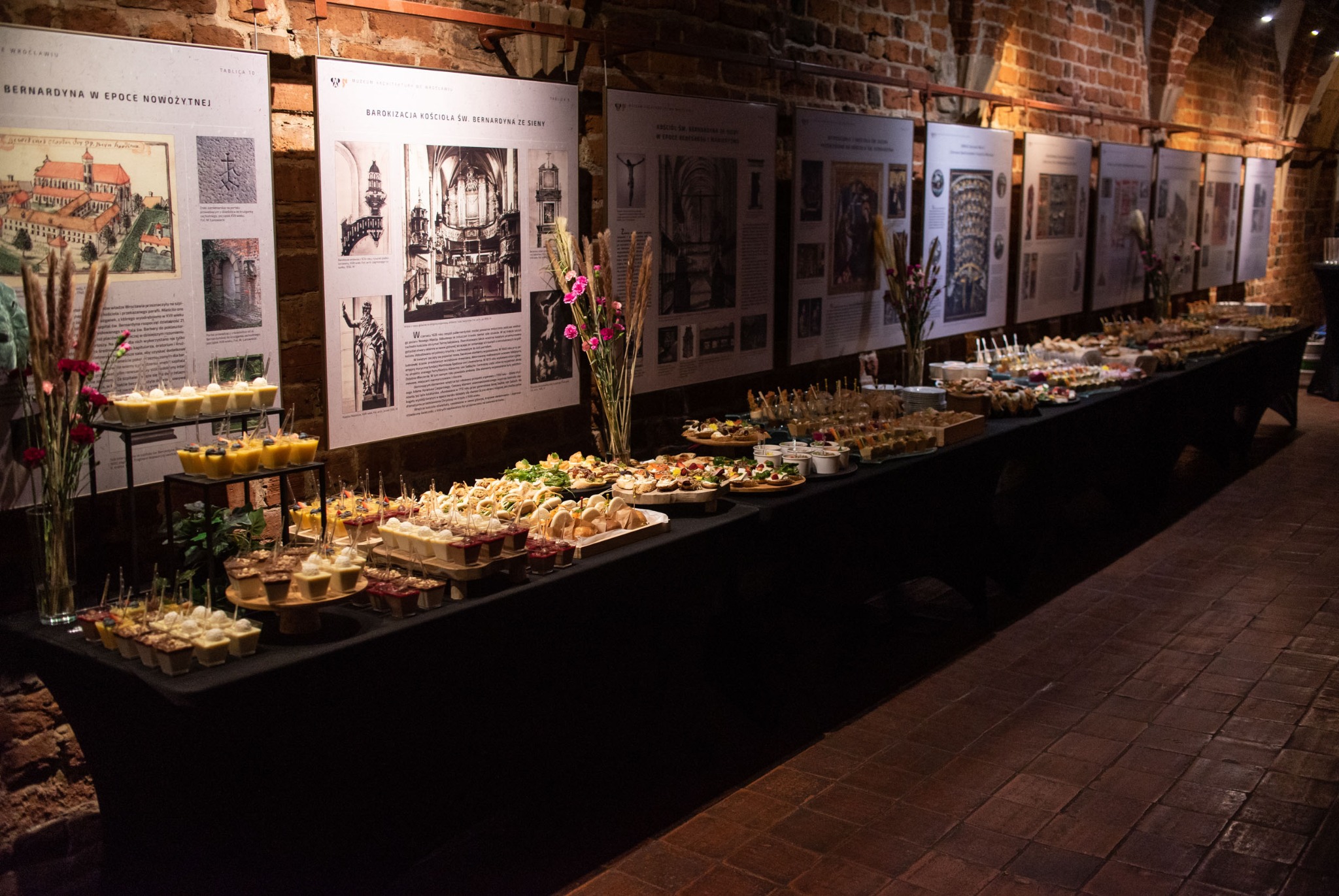 Stół cateringowy z przekąskami i deserami w zabytkowym wnętrzu z ceglanymi ścianami i planszami informacyjnymi. Różnorodne dania na czarnym obrusie, kwiaty w wazonach.