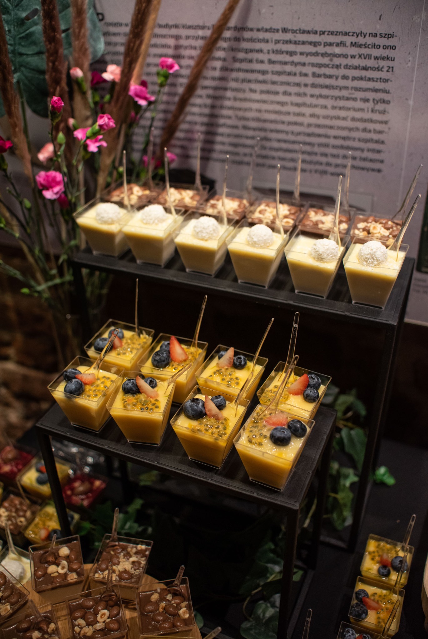 Elegancki catering: desery w małych pojemnikach, z różnymi smakami i dekoracjami, ustawione na czarnym regale. W tle kwiaty i tekst na ścianie.
